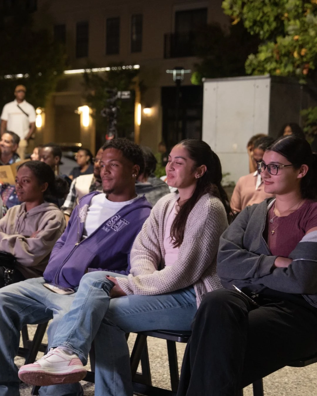 People sitting outdoors at night, some smiling, attending an event or performance.