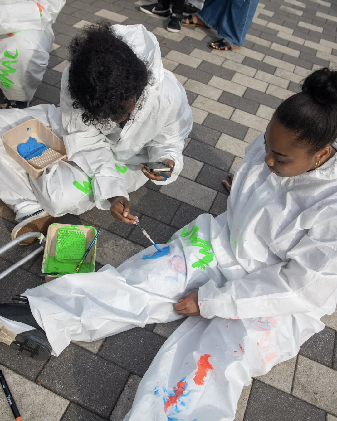 Two women in white protective suits sitting on a paved surface, painting on each other's arms with bright blue, red, and green paint, surrounded by containers of paint and brushes.