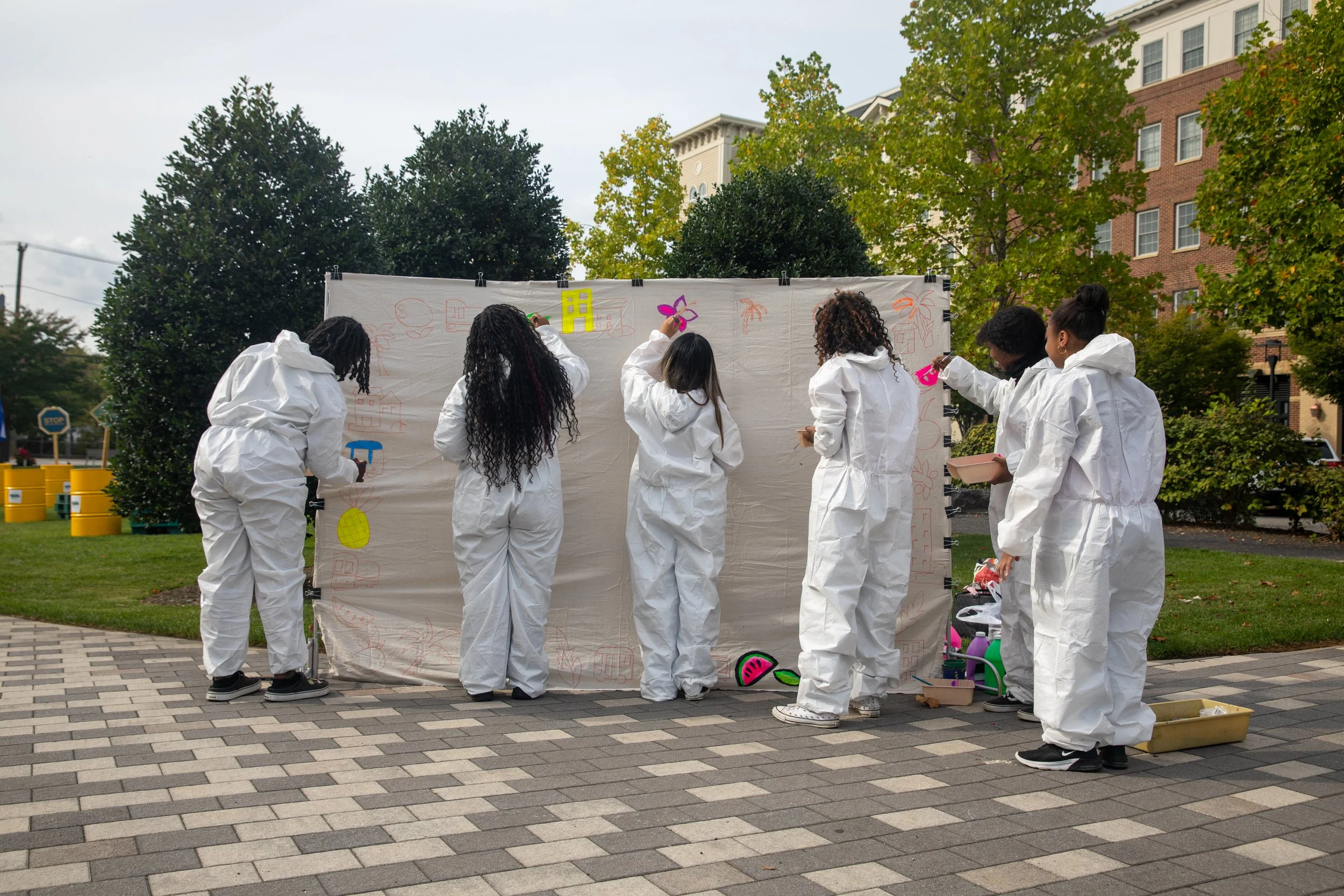 A group of people dressed in white coveralls painting a large fabric backdrop outdoors surrounded by green trees and urban buildings.