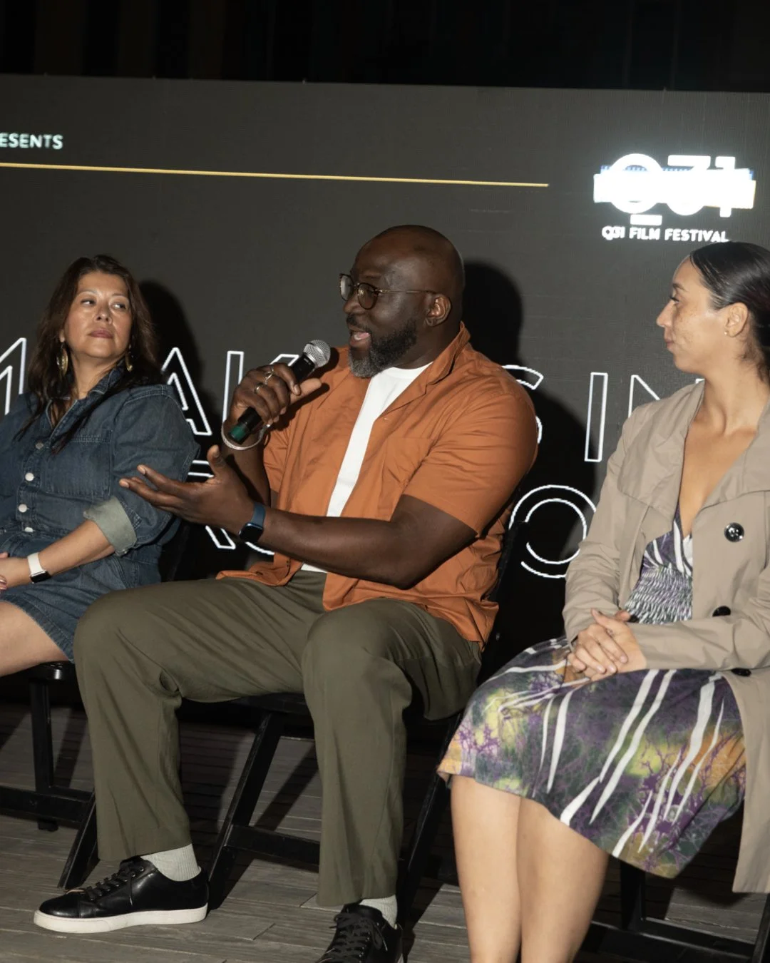 A panel discussion with three people seated on stage at OUA Film Festival. The man in the center is speaking into a microphone, flanked by two women listening.