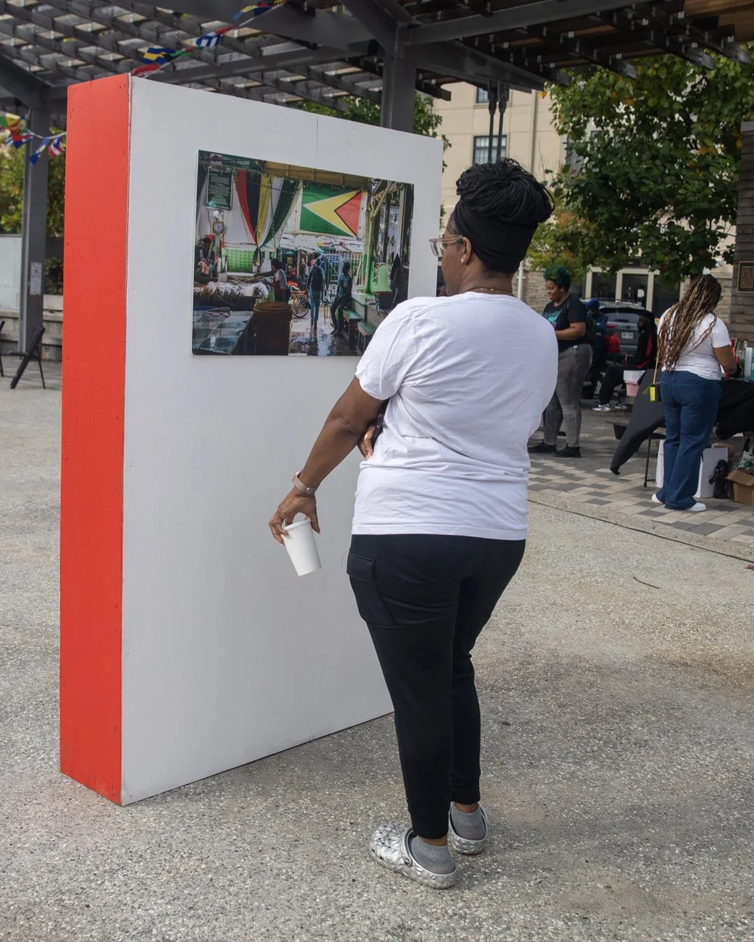 A woman in a white T-shirt, black pants, and silver shoes, with glasses and a headwrap, stands outdoors viewing a photograph on a display board at an outdoor event.