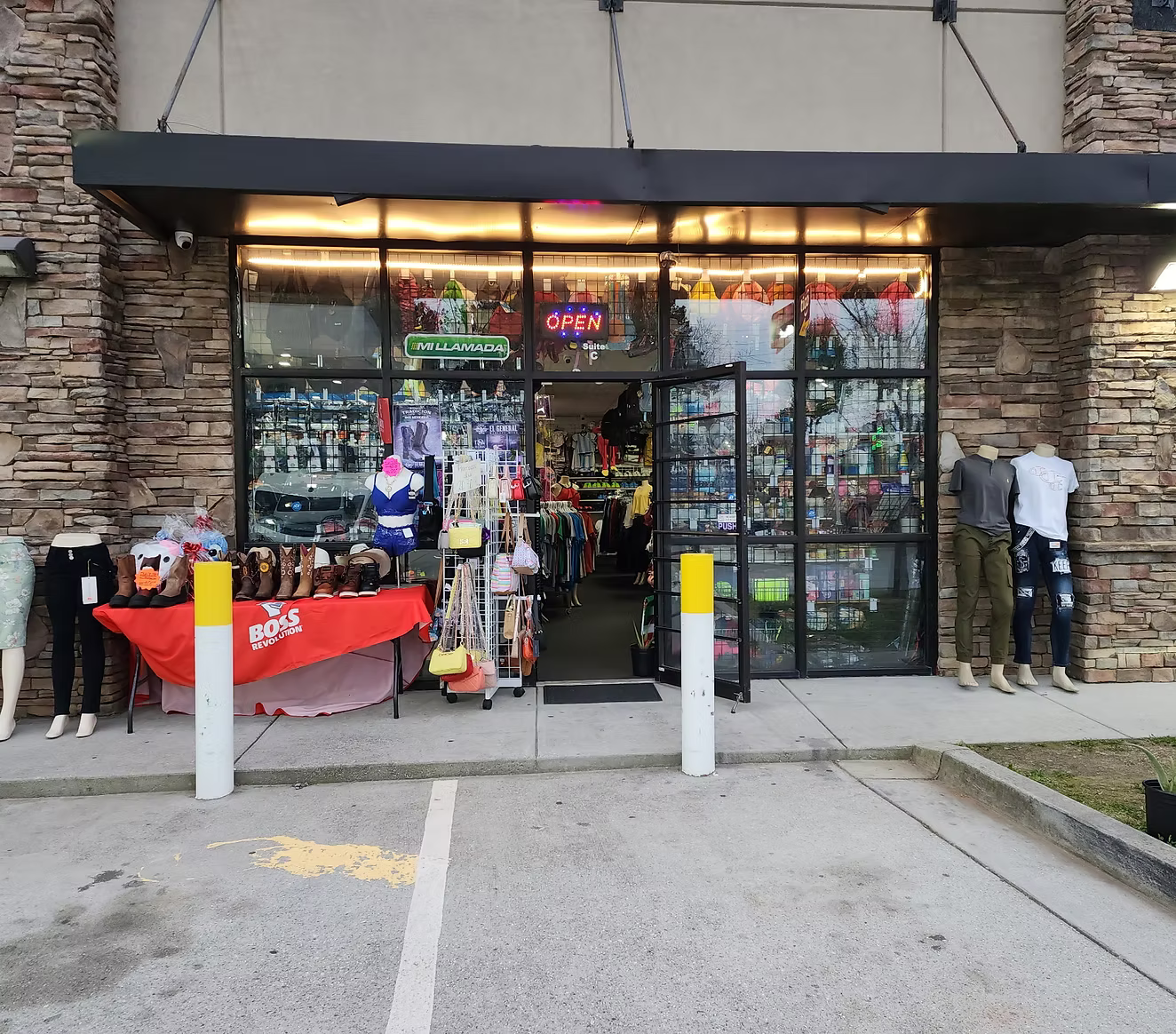 Front view of a retail store entrance with mannequins displaying clothing, a table with boots and shoes, and various accessories and apparel inside the store. The store has a neon 'OPEN' sign and glass windows that reveal more products inside.