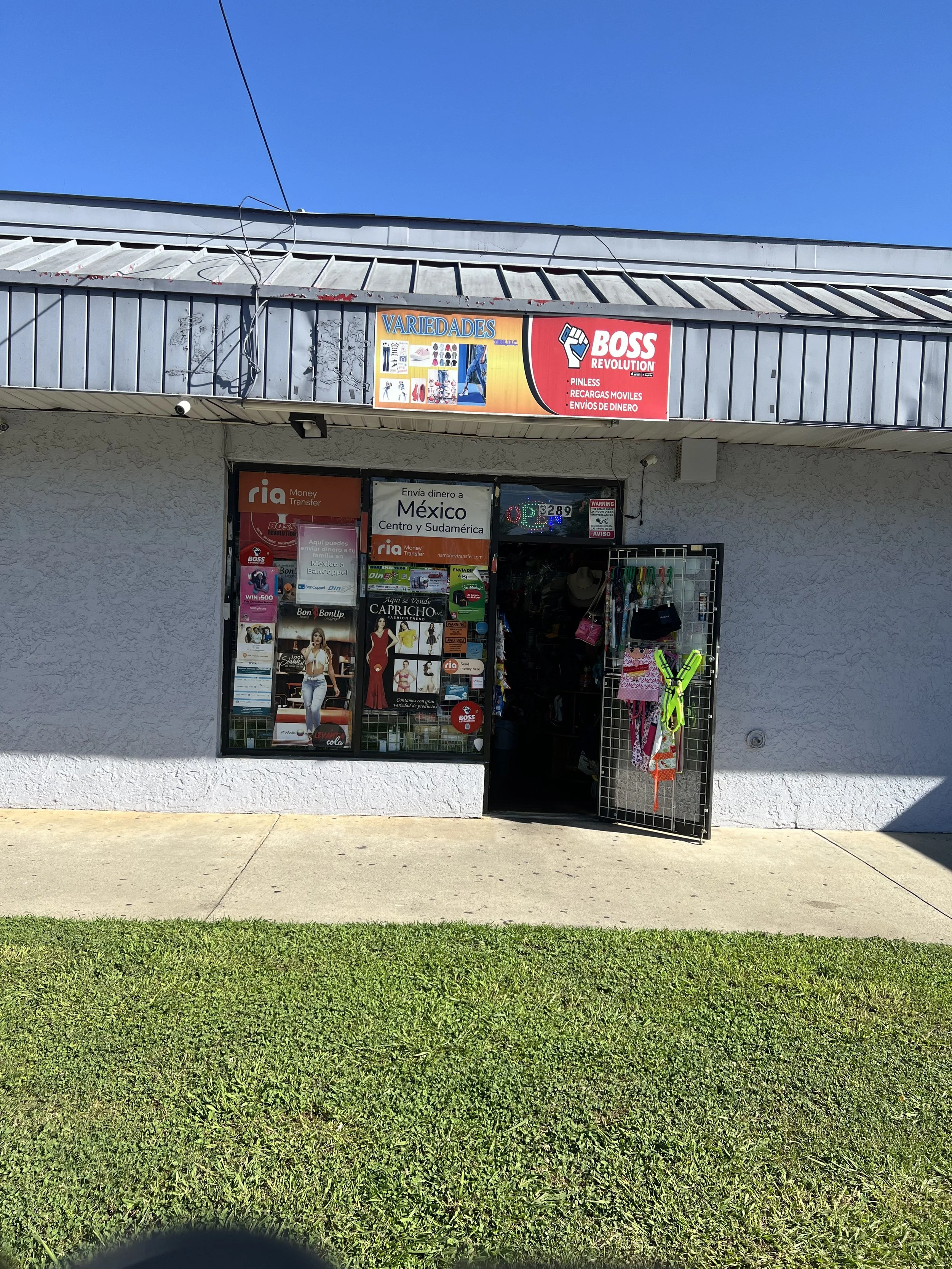 The storefront of a small shop with signs advertising money transfer services to Mexico and a variety of other products, with clothing and accessories displayed outside, under a clear blue sky.