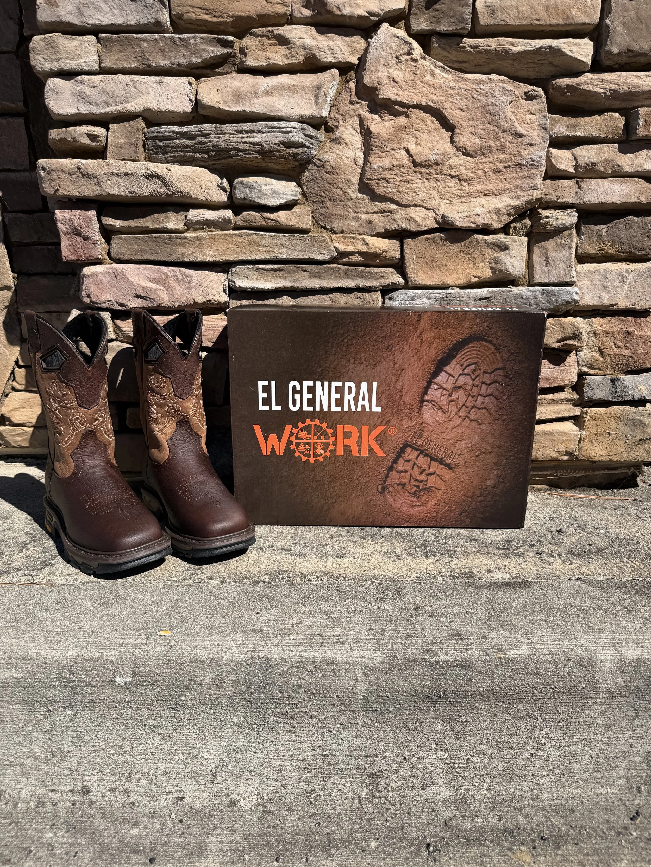 Pair of brown cowboy boots, a box with the logo 'EL GENERAL WORK', and a stone wall background.