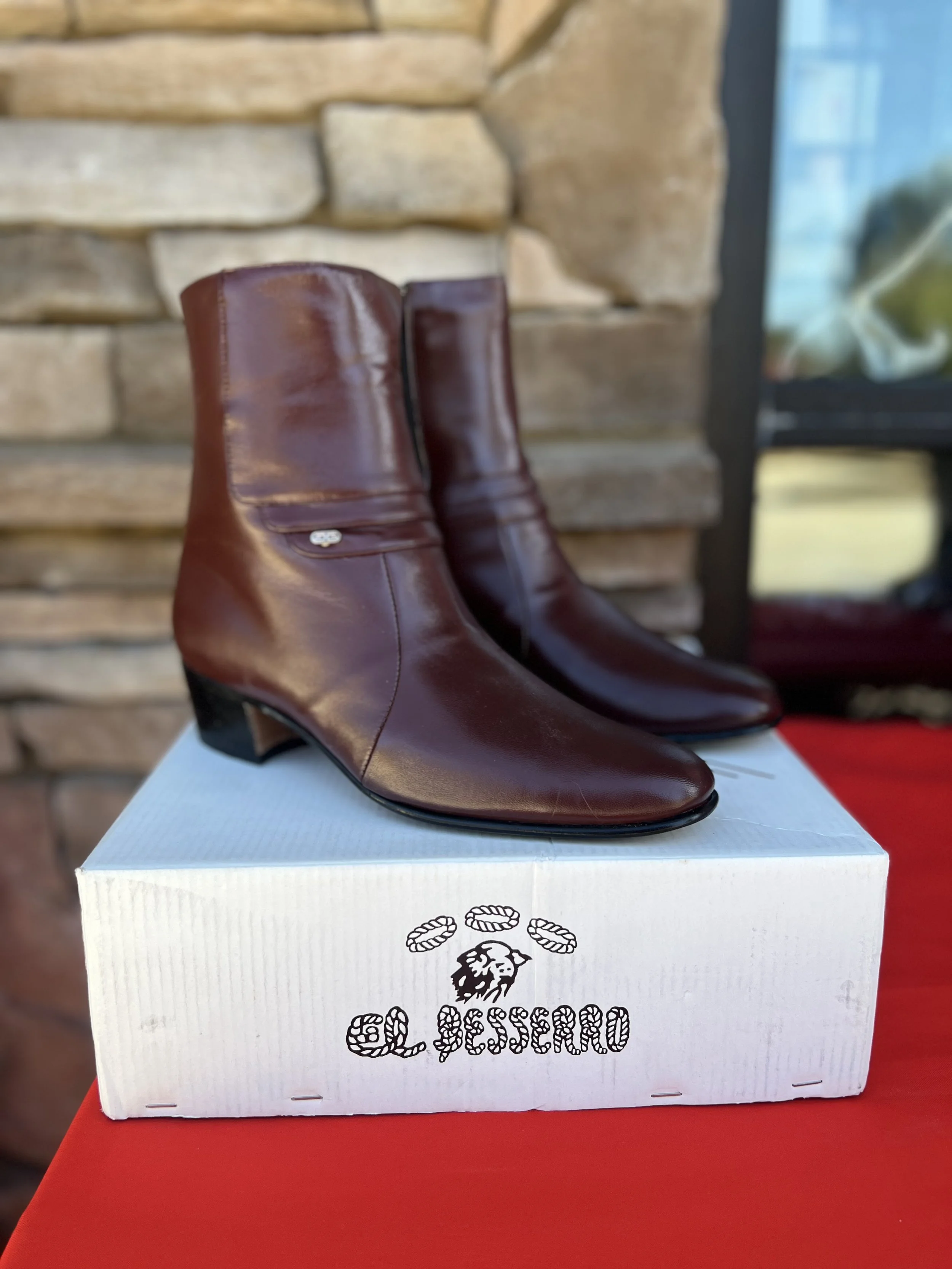 A pair of shiny brown leather cowboy boots positioned on a white box with a logo and the text "GLESSARO." The box is placed on a red surface, with a stone and brick wall in the background.