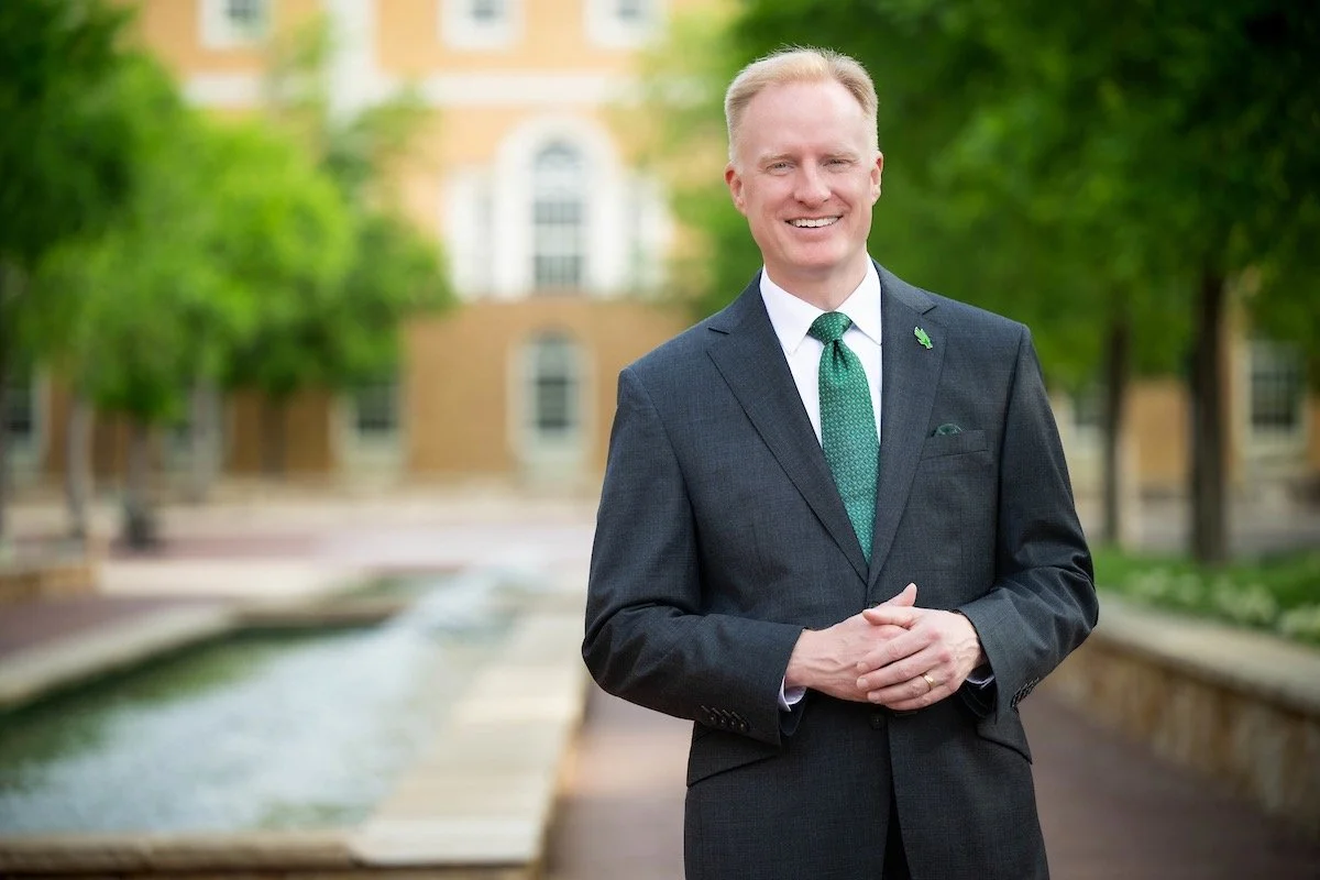 President Harrison Keller of UNT in a dark suit, white shirt, and green tie standing outdoors in front of the Hurley Administration Building with trees, smiling with hands clasped.