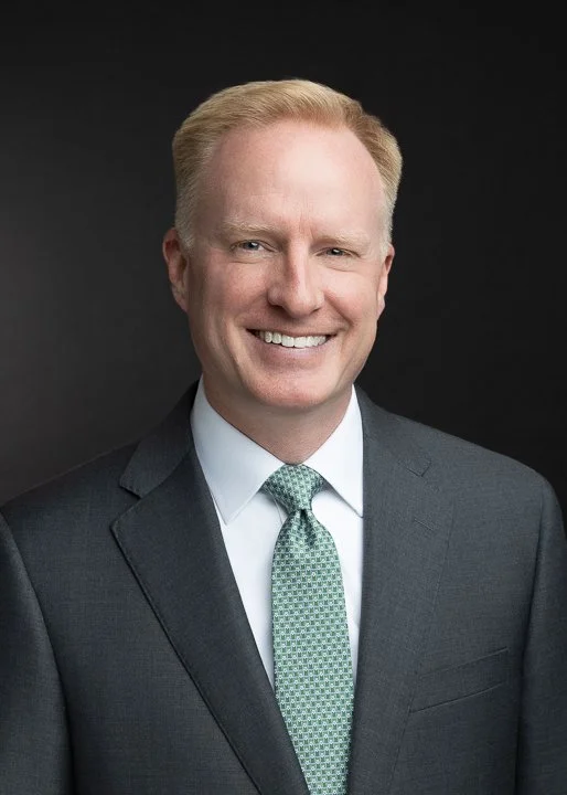 Professional headshot of a man in a dark gray suit, white shirt, and green patterned tie, smiling against a dark background.