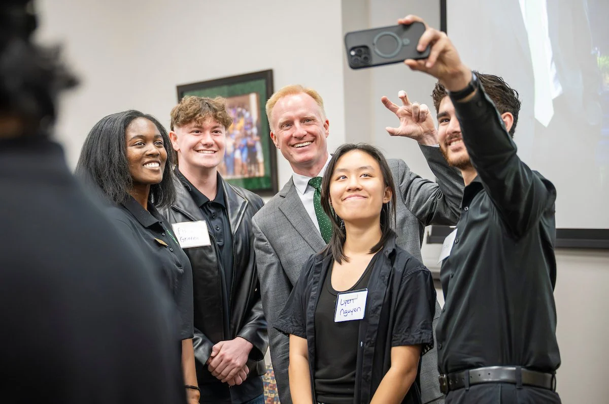 Group of five UNT students and President Harrison Keller taking a selfie together at an indoor event, smiling and posing for the camera.