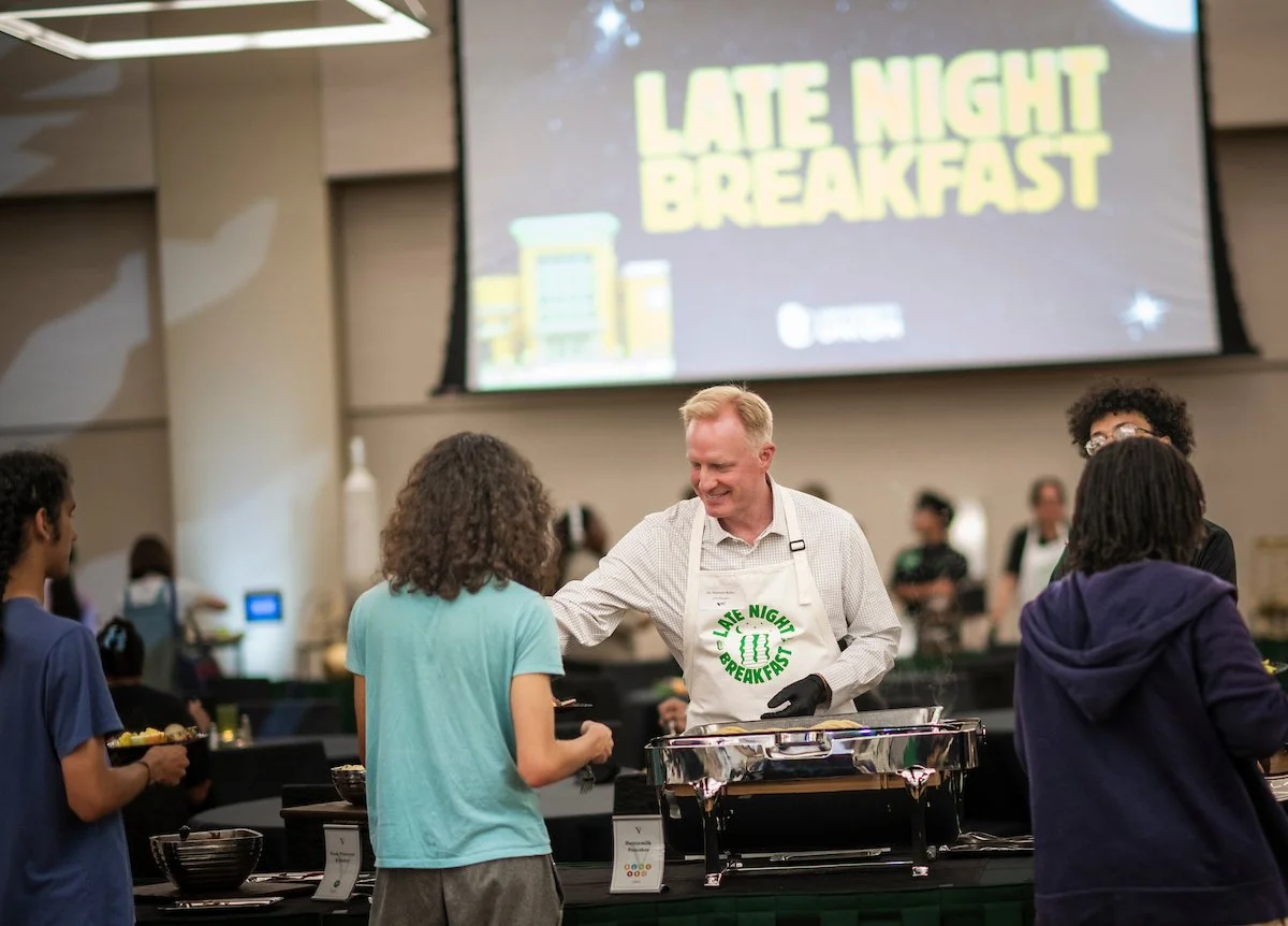 President Harrison Keller with a white shirt and apron serving food at the 'Late Night Breakfast' event in a large room at the UNT Student Union, with several students lined up with plates.