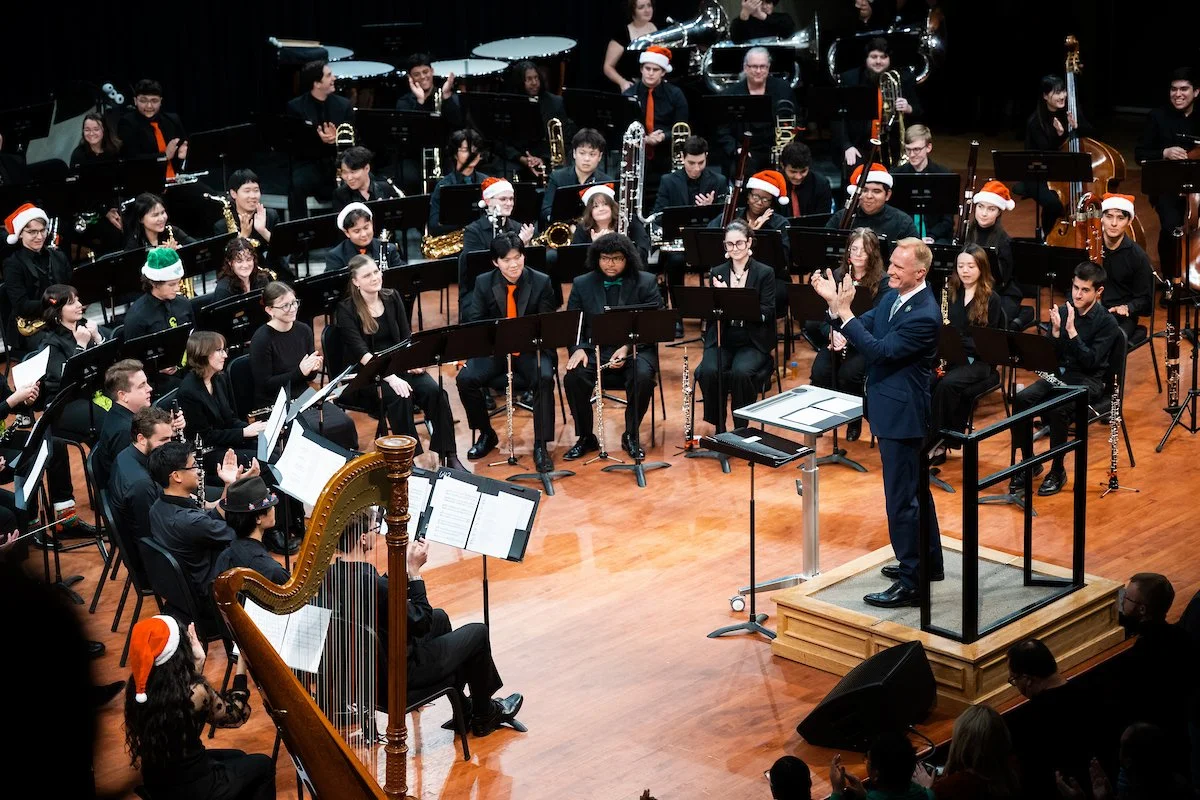 The UNT wind orchestra performing on stage with President Harrison Keller on the podium, many members wearing Santa hats and Christmas accessories, with a musician playing a harp in the foreground.