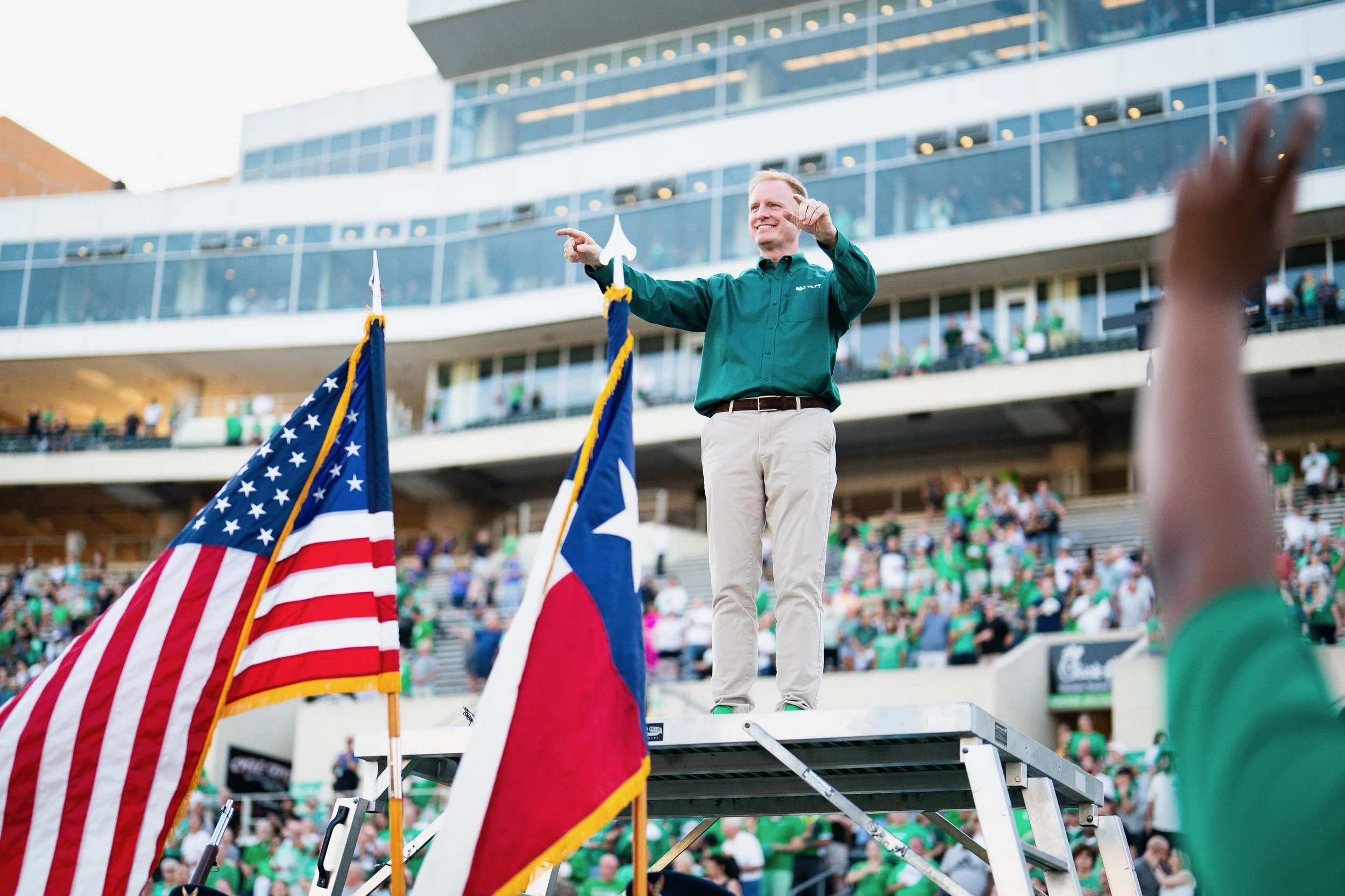 President Keller of UNT standing on a podium, smiling and directing the Green Brigade marching band during a home football game. Flags are visible in the foreground, with a crowd watching in the background.