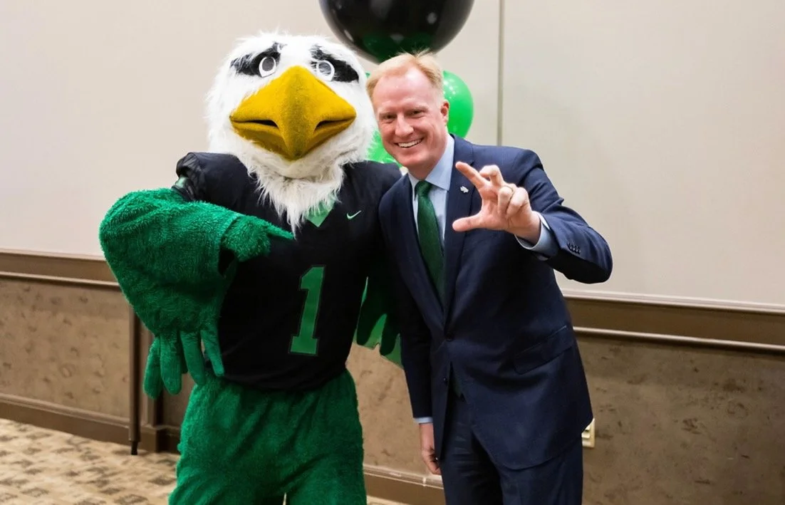 President Keller of UNT in a suit and tie posing with the UNT mascot Scrappy the Eagle, both smiling at the camera.