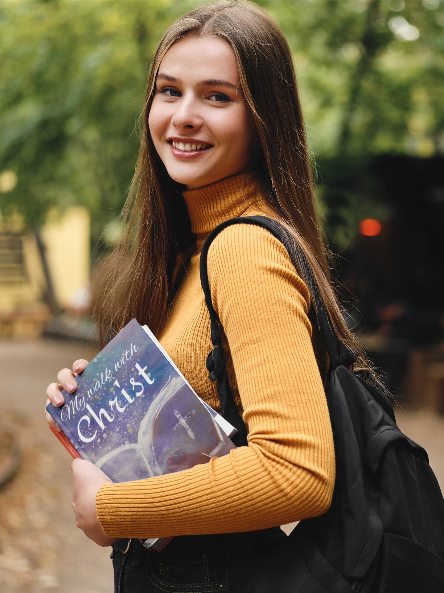 A young woman with long brown hair smiling and holding a book titled 'My walk with Christ' while standing outdoors with green trees in the background.