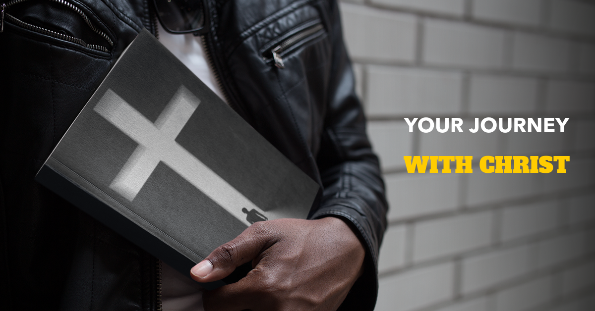 Person holding a black and white journal with a cross on the cover, standing against a brick wall.