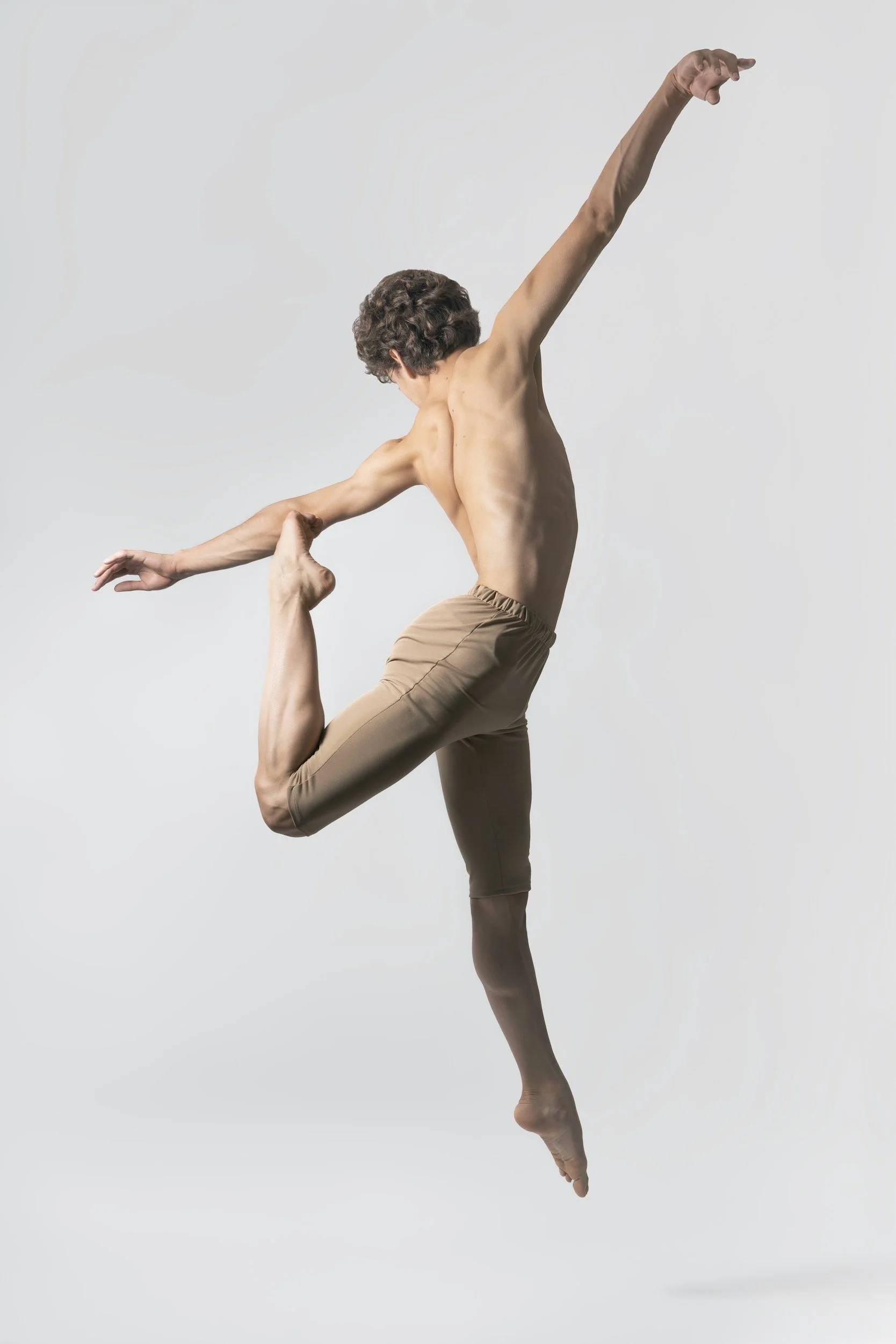 A shirtless male dancer with curly hair performing a ballet pose, holding his left ankle with his left hand while extending his right arm upward, standing on his right leg against a plain white background.