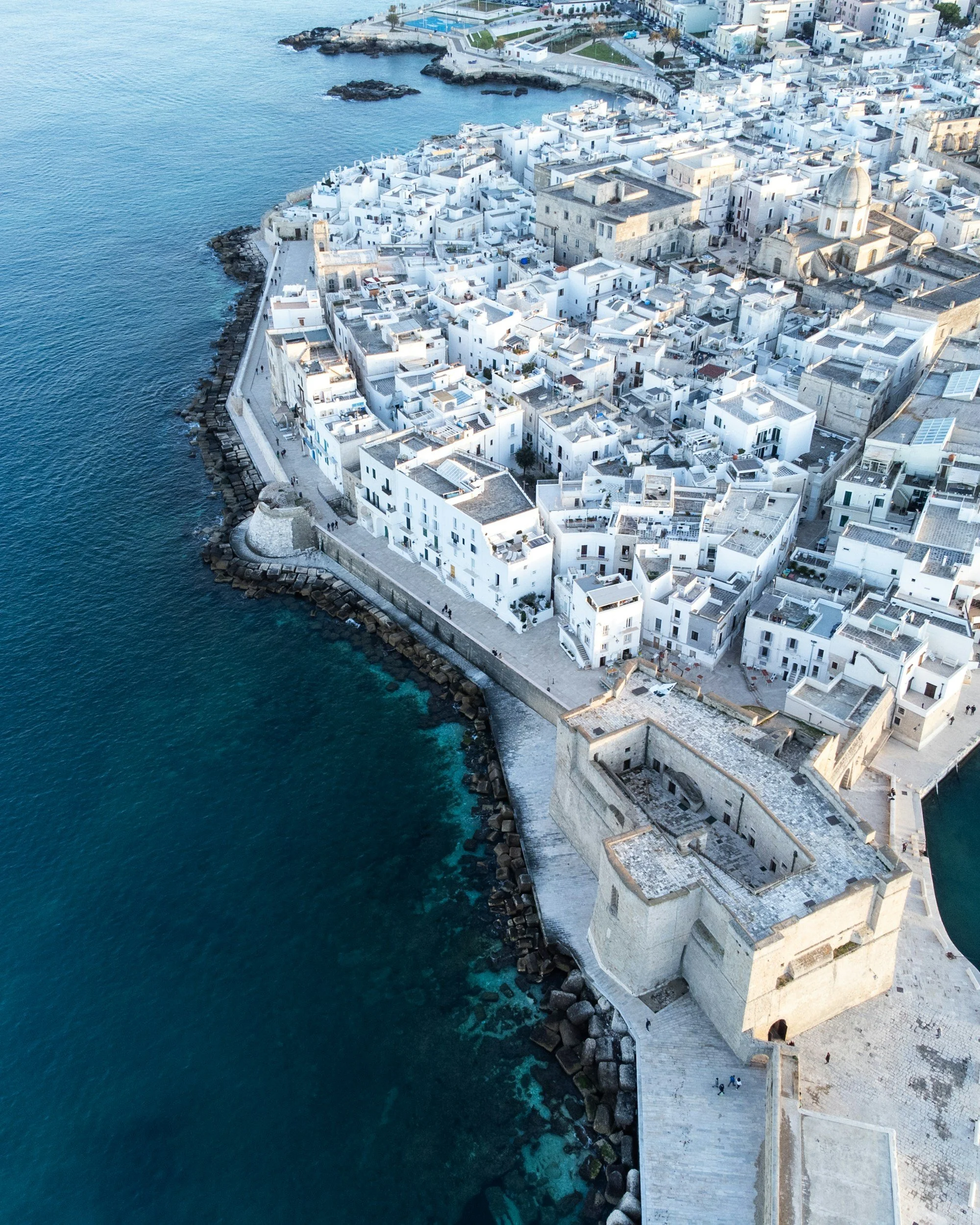 Aerial view of a historic coastal town with white buildings, a stone fortress wall along the shoreline, and turquoise ocean water.