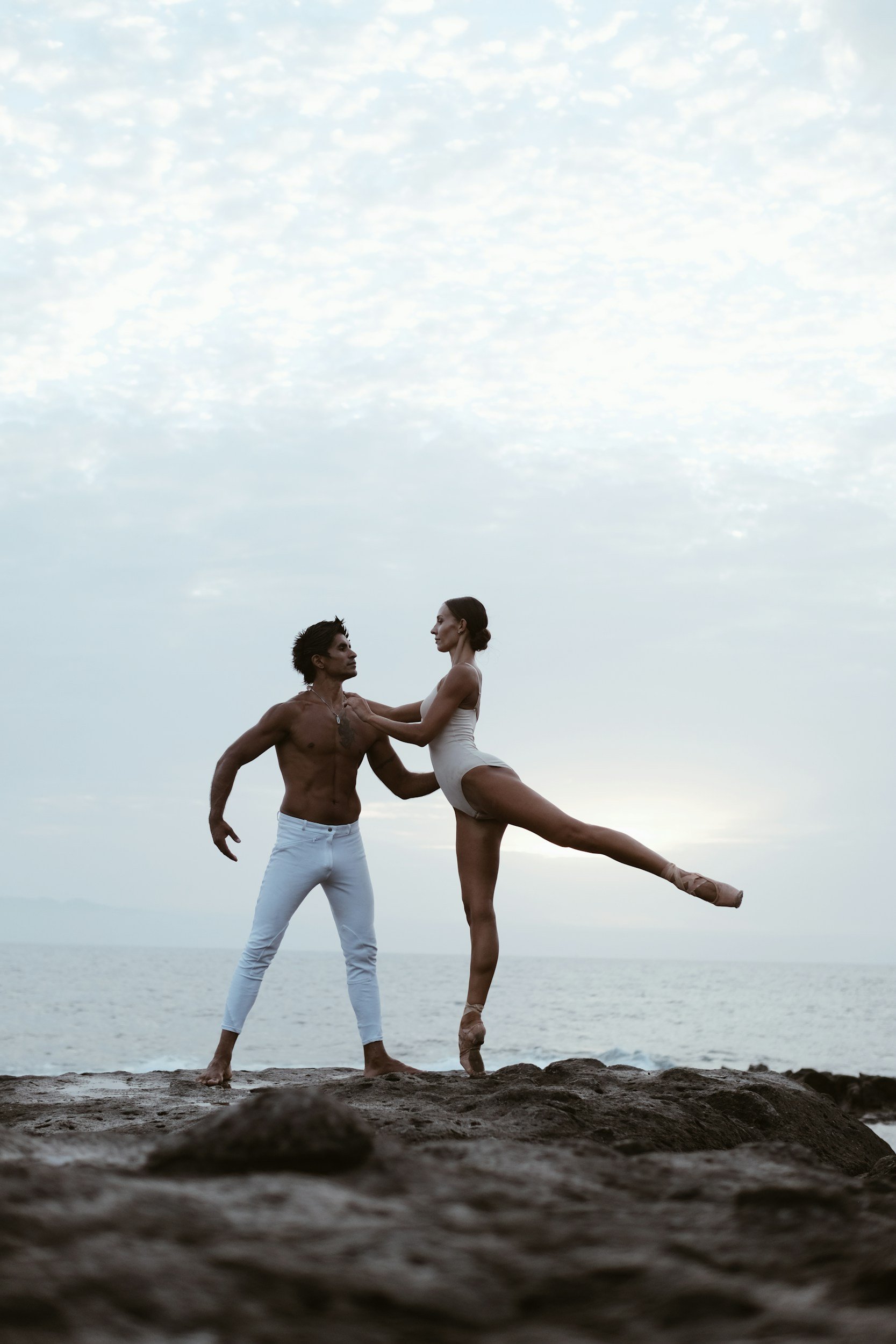 A male ballet dancer supporting a female ballet dancer in an arabesque pose on a beach at sunset or dusk.
