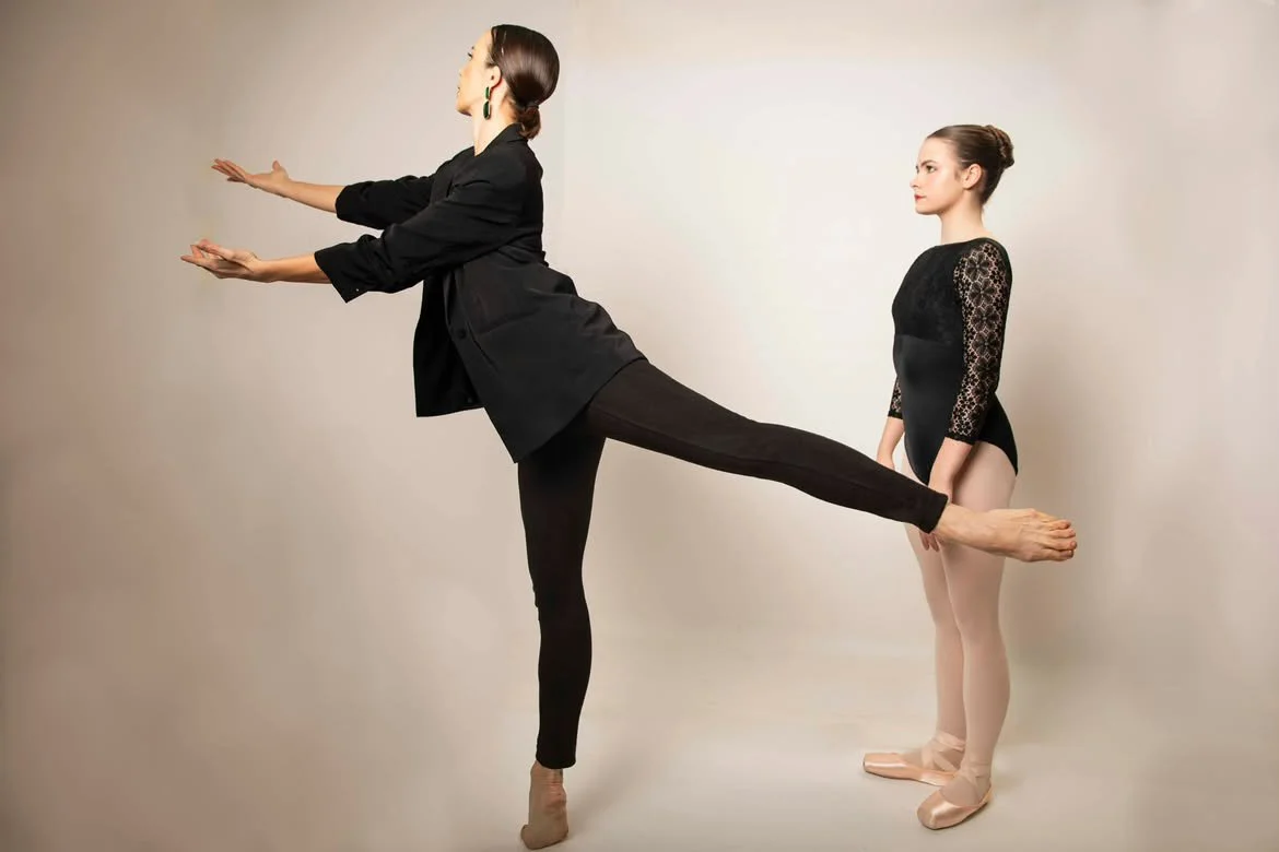 Two women in ballet attire, one in black with a blazer and the other in a black leotard with lace sleeves, participate in a ballet practice where one balances on one leg with the other leg extended behind, supported by the first woman.