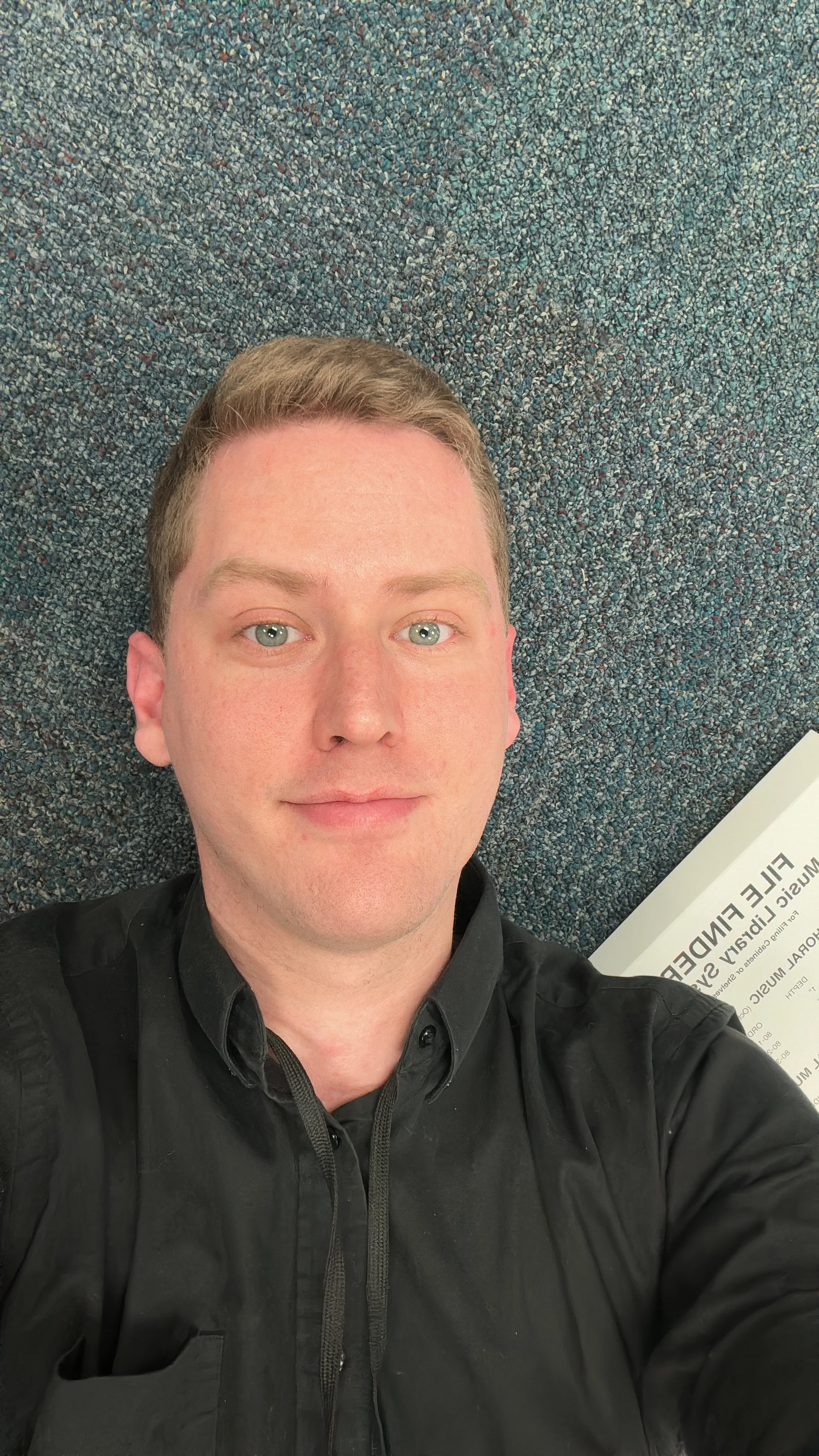 A young man with short blond hair and blue eyes taking a selfie while lying on a blue speckled carpeted floor. Part of a paper or newspaper is visible in the corner of the image.
