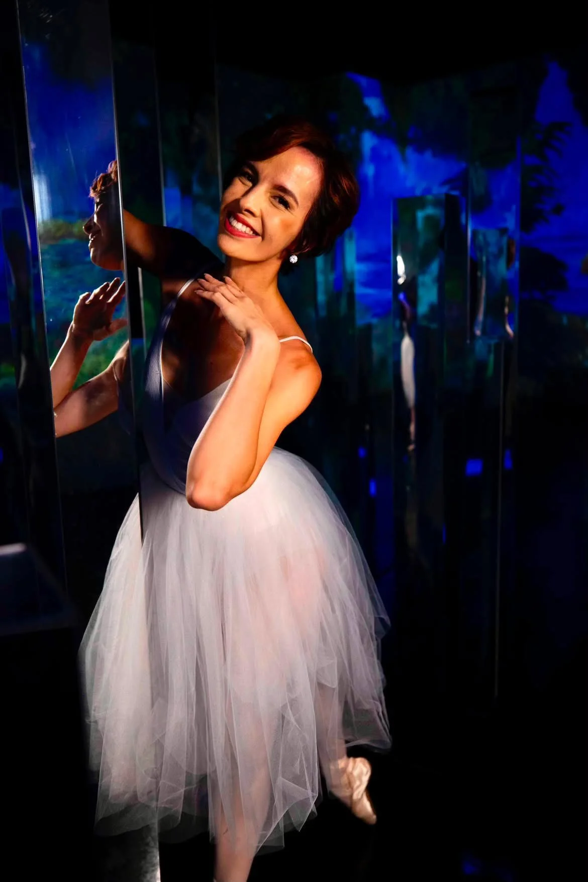 A woman in a ballet tutu smiling and posing with her hand on her shoulder, standing among mirrors in a dark room with blue lighting.