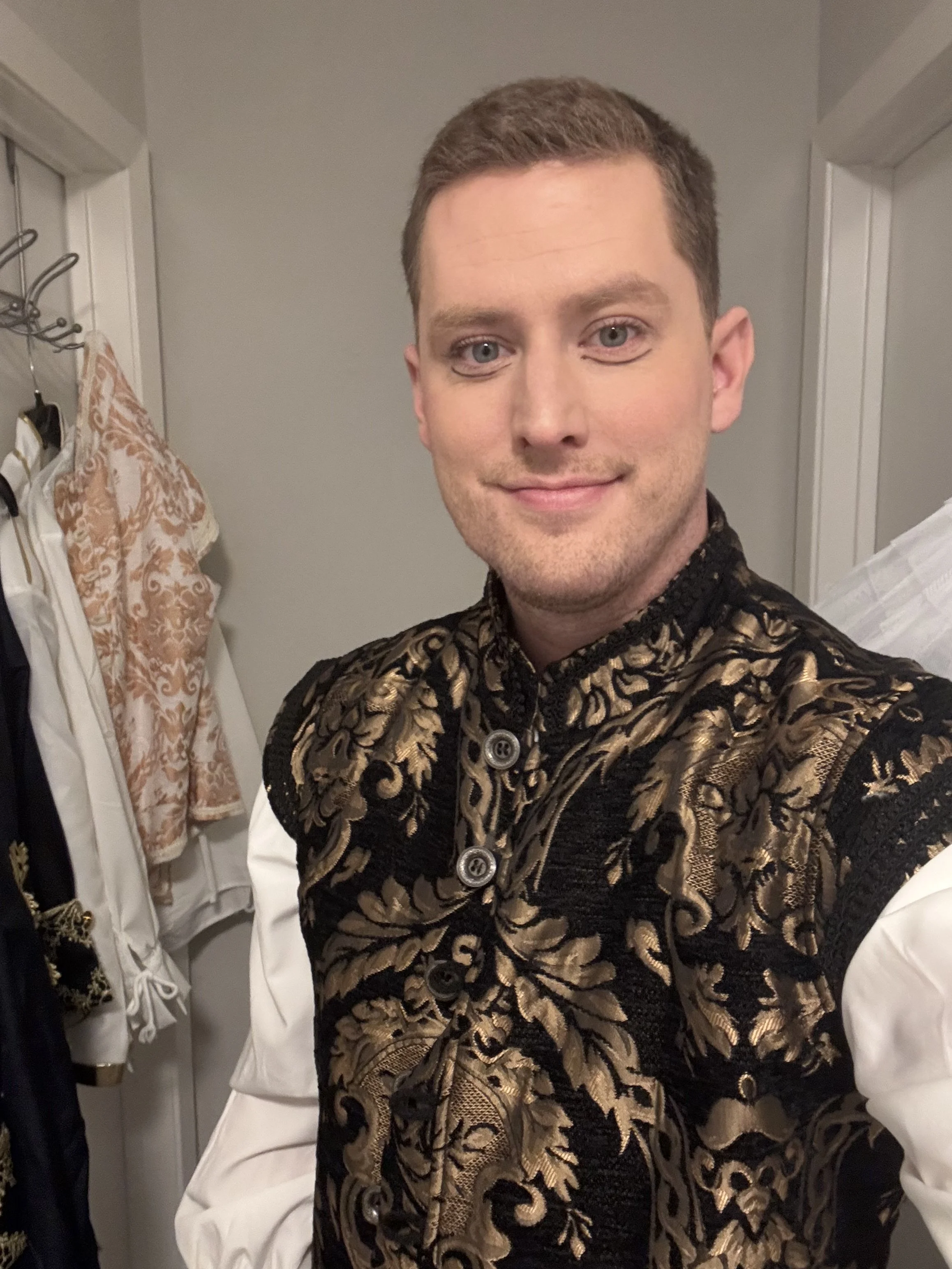 A young man with short brown hair and blue eyes taking a selfie in a mirror, wearing a black and gold ornate patterned jacket, standing in a room with clothing hanging on hooks and a gray wall in the background.