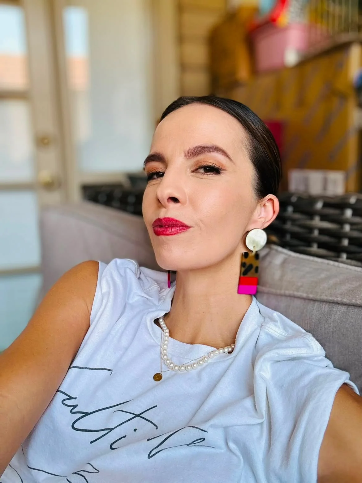 A woman with dark hair and bright lipstick taking a selfie indoors, wearing statement earrings, a pearl necklace, and a white T-shirt with black cursive writing.