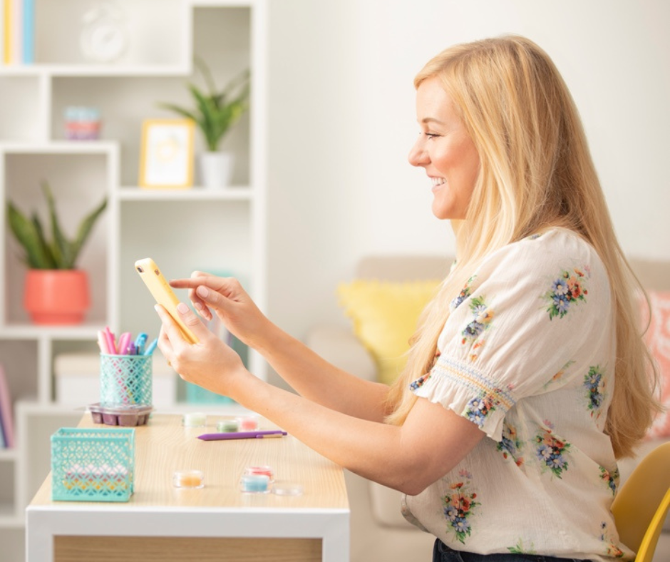 A woman with blonde hair and a floral blouse smiling and looking at her phone in a brightly decorated room with white shelves, plants, and colorful stationery. She is thinking about what to add to her Scentsy subscription and what item she will recei