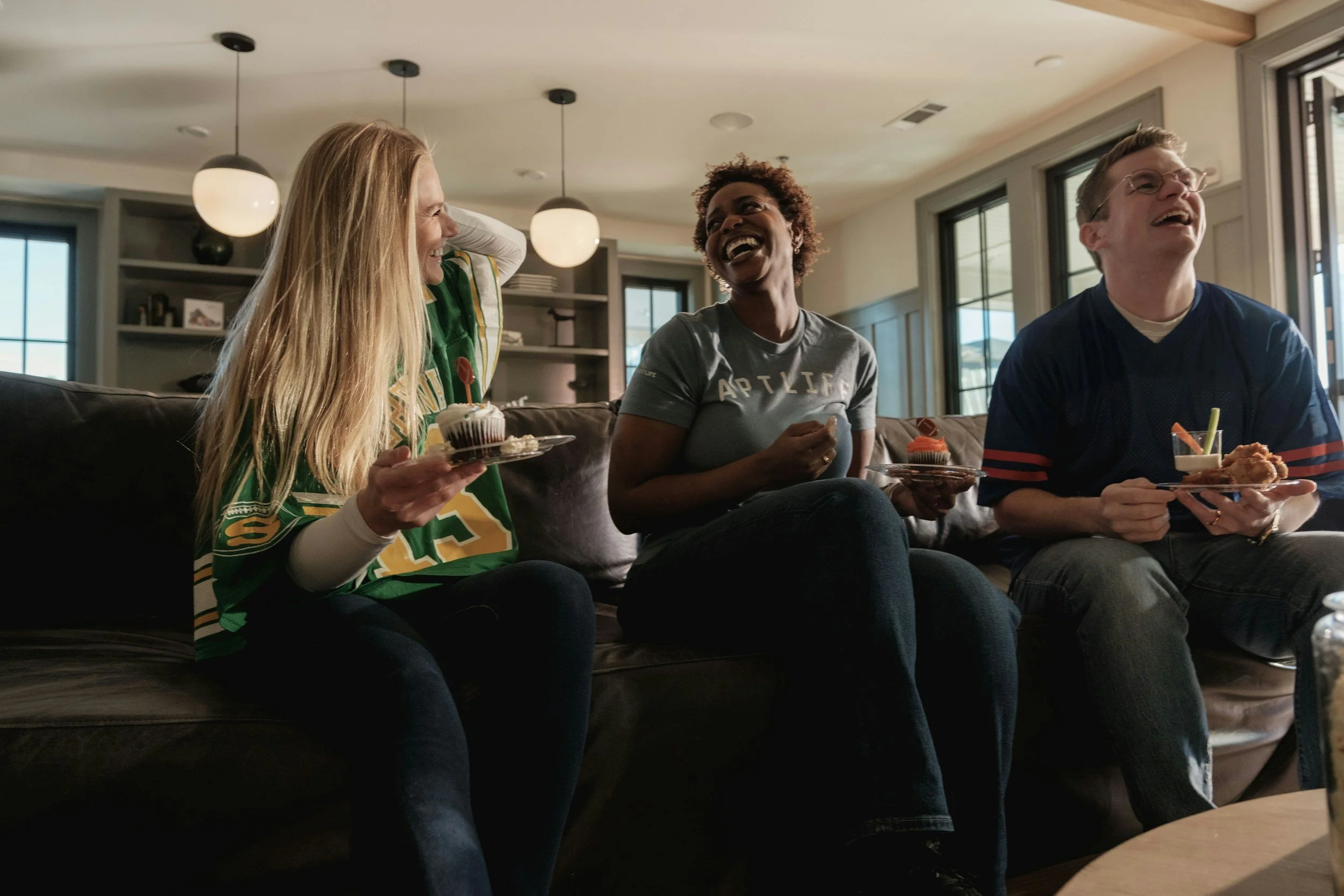 Three friends sitting on a couch, laughing and enjoying snacks in a bright room with large windows, hanging pendant lights, and bookshelves. They have gathered for a Scentsy fragrance event.