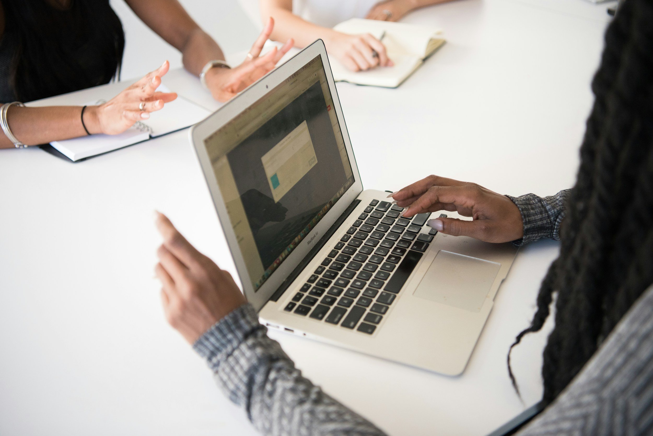Close-up of a woman using a MacBook laptop at a white table during a meeting with other people. Her hands are on the keyboard, and she is wearing a plaid shirt.