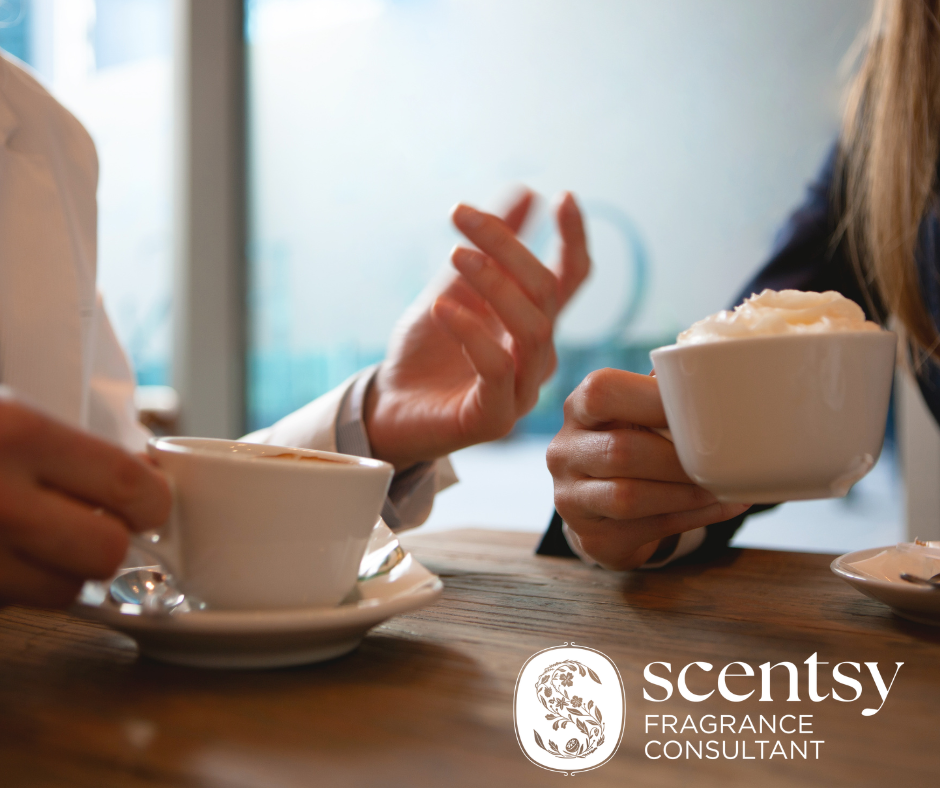 Two people sitting at a wooden table, each holding a cup of coffee. They are chatting about different event options Scentsy has to offer.
