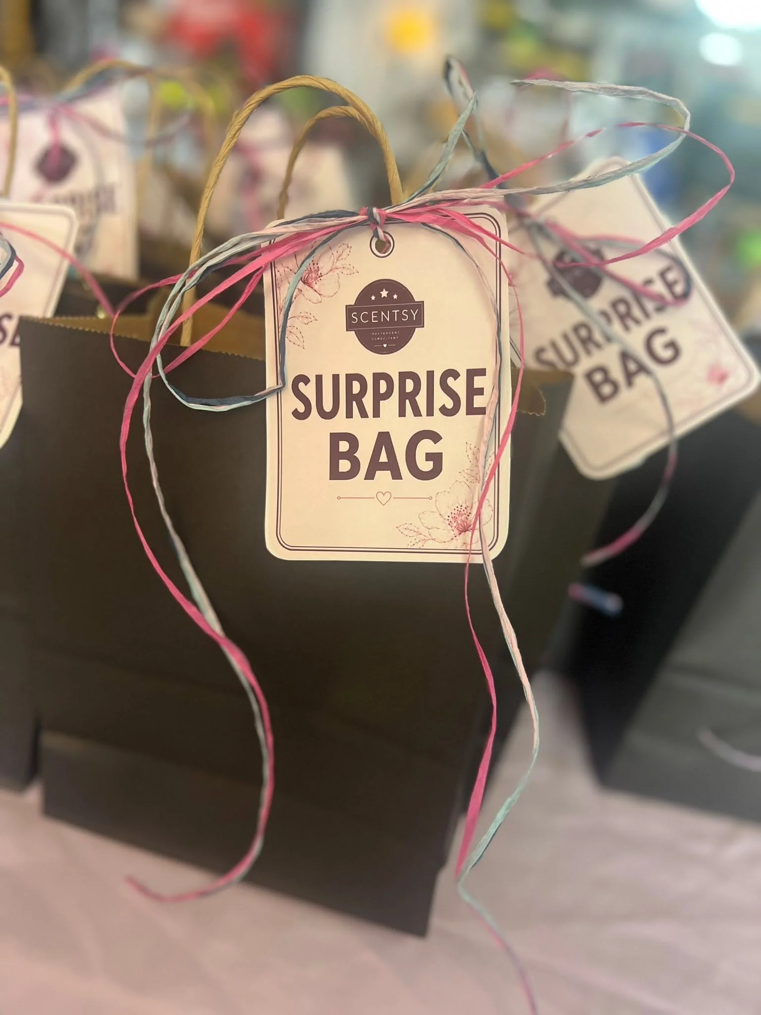 Small paper gift bags labeled 'Surprise Bag' with colorful ribbons tied on top, displayed on a shelf or counter. The bags are waiting to be given to Scentsy party guests.