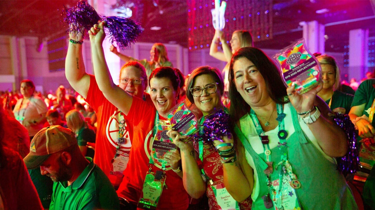 Group of smiling women celebrating each other at Scentsy Family reunion 2025, holding colorful reflective materials, wearing lanyards and accessories, with a lively crowd and stage lighting in the background.