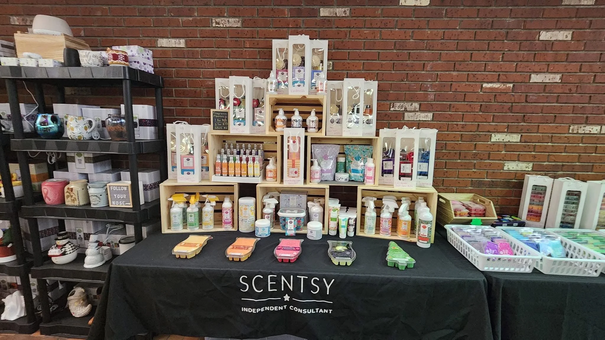 Craft fair display of scented products, hand sanitizers, and small gift items on wooden and black shelves, with a table covered by a black cloth labeled 'SCENTSY' in front, against a brick wall background.