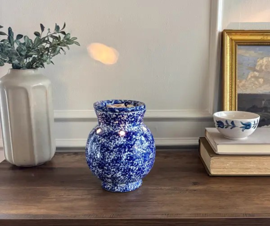 A wooden mantel with a large off-white vase containing greenery, a Blue Spongeware Scentsy Warmer, a white bowl with blue floral design, and a framed painting.