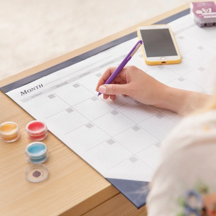 A person writing the date of their Scentsy Club shipment on a blank monthly planner with a purple pen, surrounded by colorful paints, a smartphone, and a pink eraser on a wooden desk.