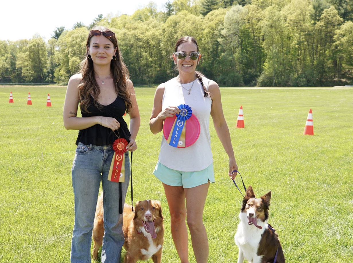 Two women standing on a green field with their dogs, holding ribbons for dog competition awards, with orange cones in the background.
