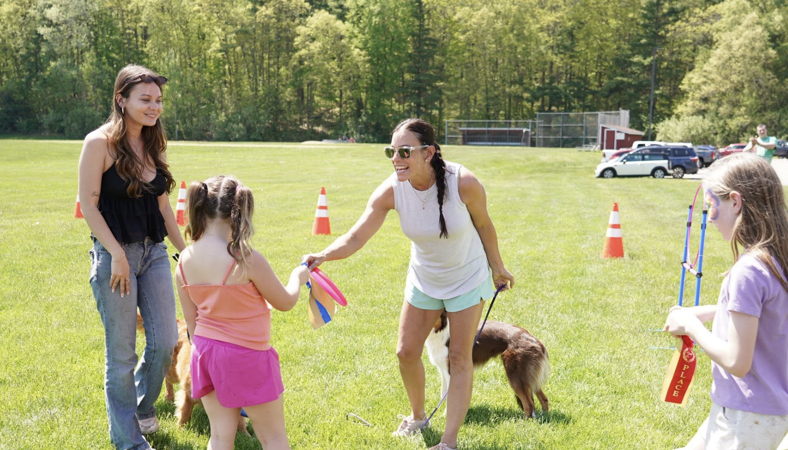 A woman handing a ribbon to a young girl during a dog competition in a grassy field on a sunny day, with other girls, a woman, and parked cars visible in the background.
