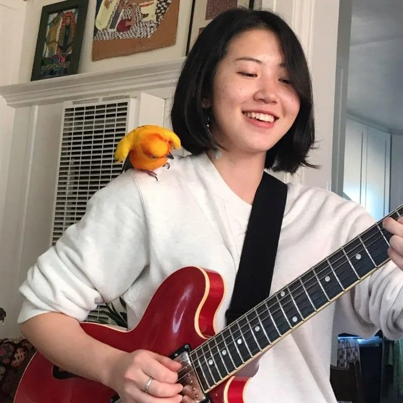 A young woman with shoulder-length black hair and light skin, smiling and playing an electric guitar, with a colorful yellow and orange bird perched on her shoulder.
