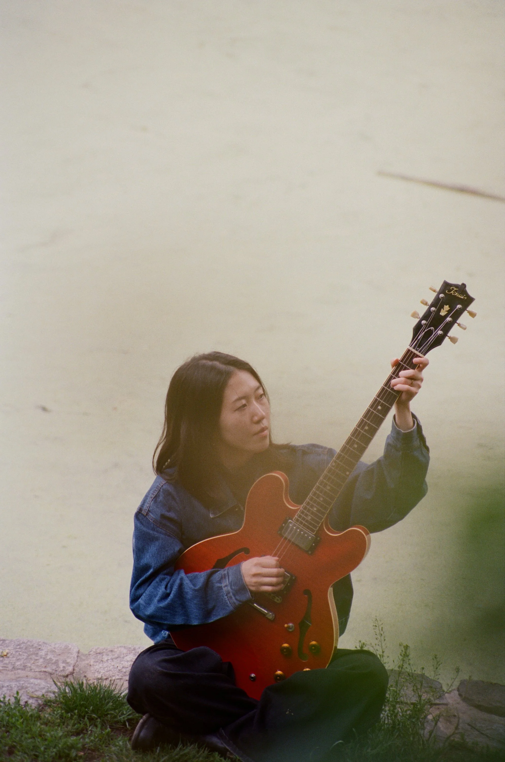 A young woman sitting outside on the grass, holding a red hollow-bodied electric guitar. She is wearing a denim jacket and black pants, with her long dark hair worn down. The background features a plain, light-colored wall.