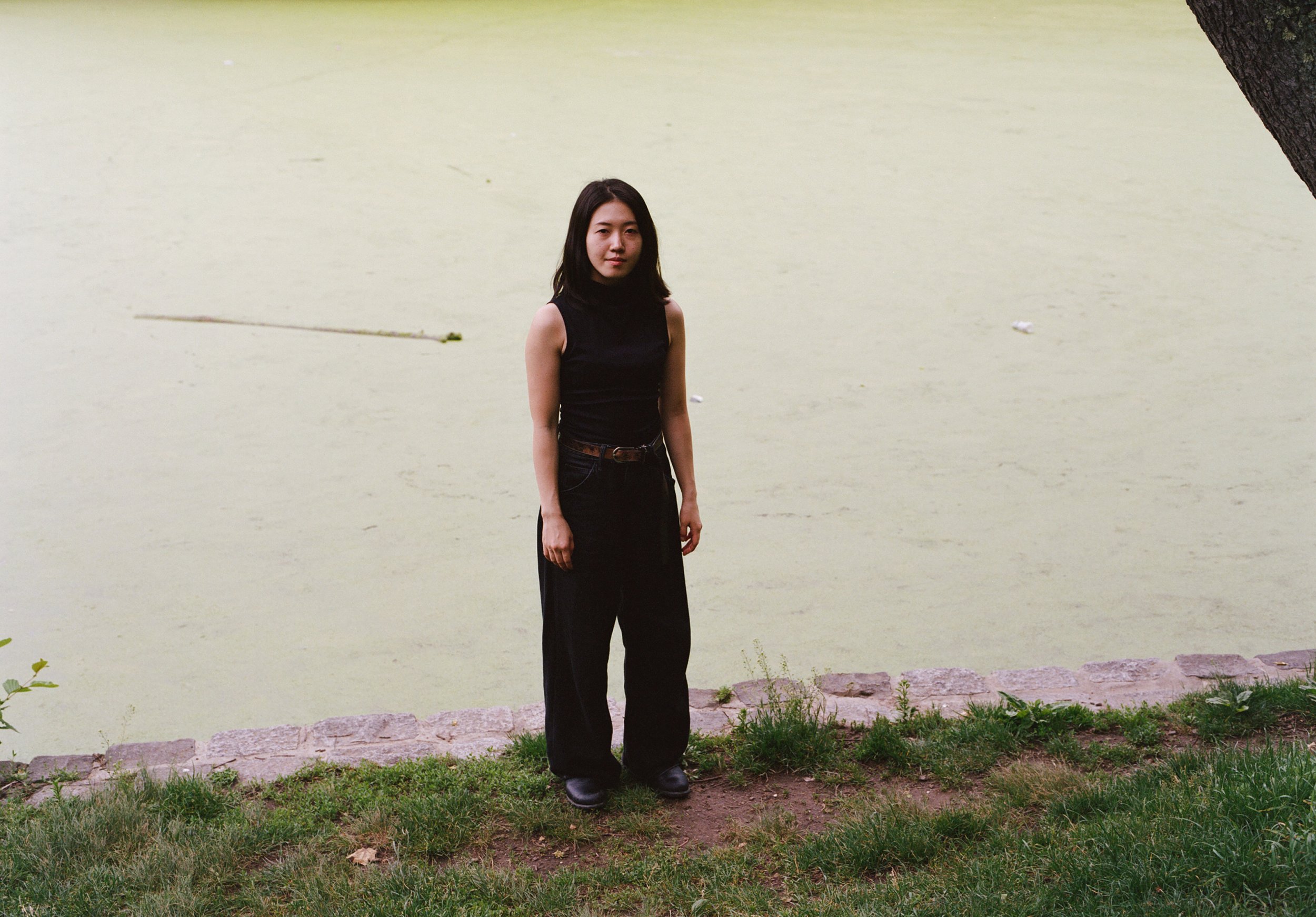 A woman standing near a lake covered with green duckweed, with a large tree trunk on the upper right corner and some grass and rocks at the ground.
