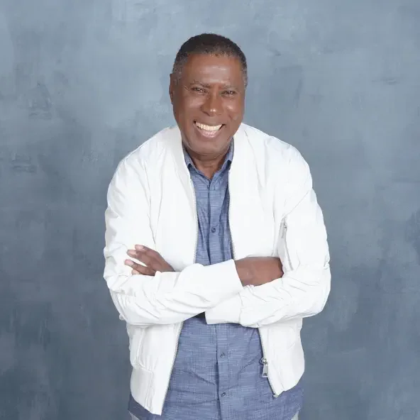 A smiling man with crossed arms wearing a white jacket and blue shirt against a gray background.
