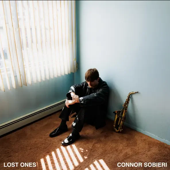A young man sitting on the brown carpeted floor, leaning against a light blue wall in a corner, with a gold saxophone resting against the wall next to him. Sunlight streams through vertical blinds, casting striped shadows on the floor.