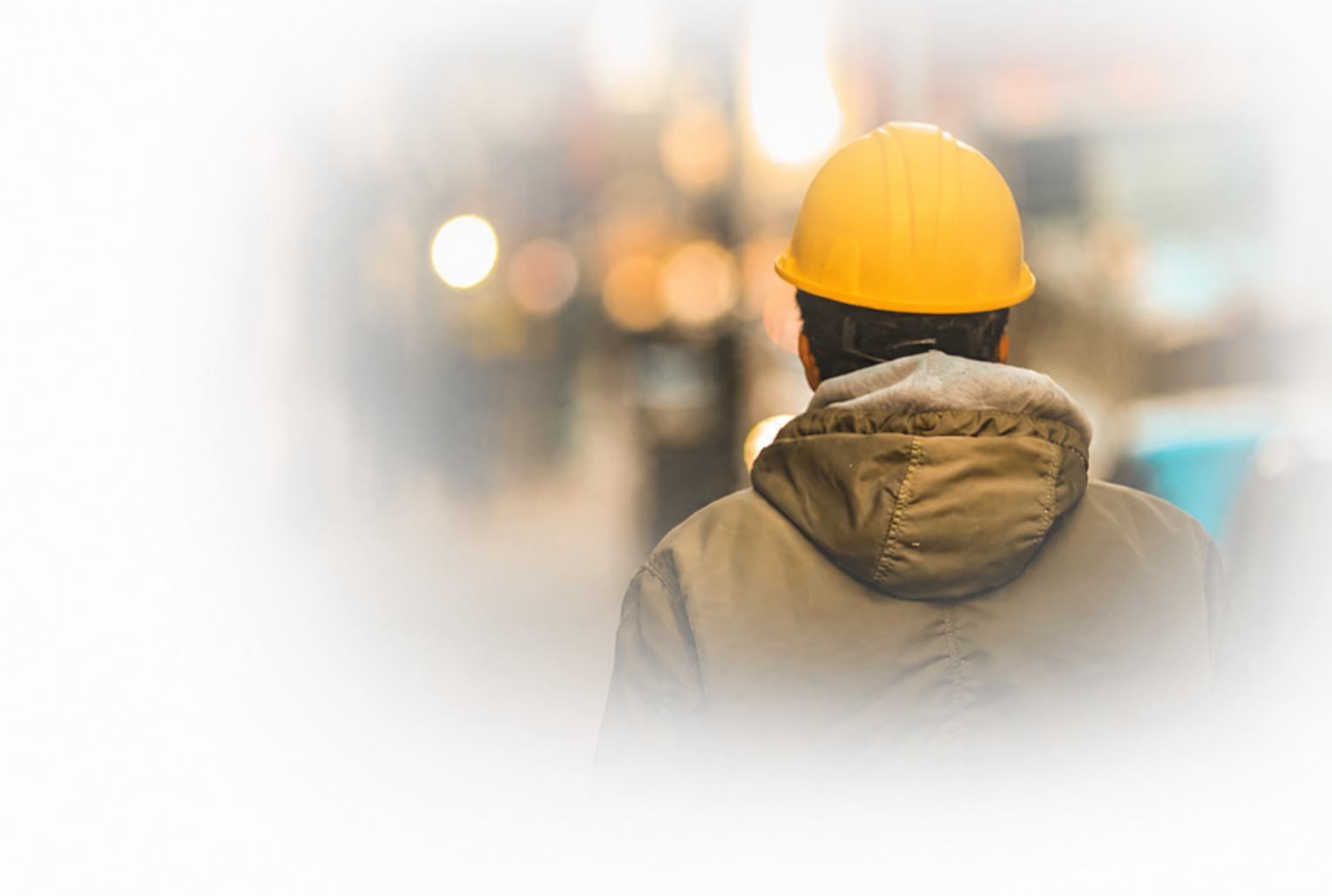 Back view of a construction worker wearing a yellow safety helmet and a brown jacket in an urban setting with blurred lights in the background.