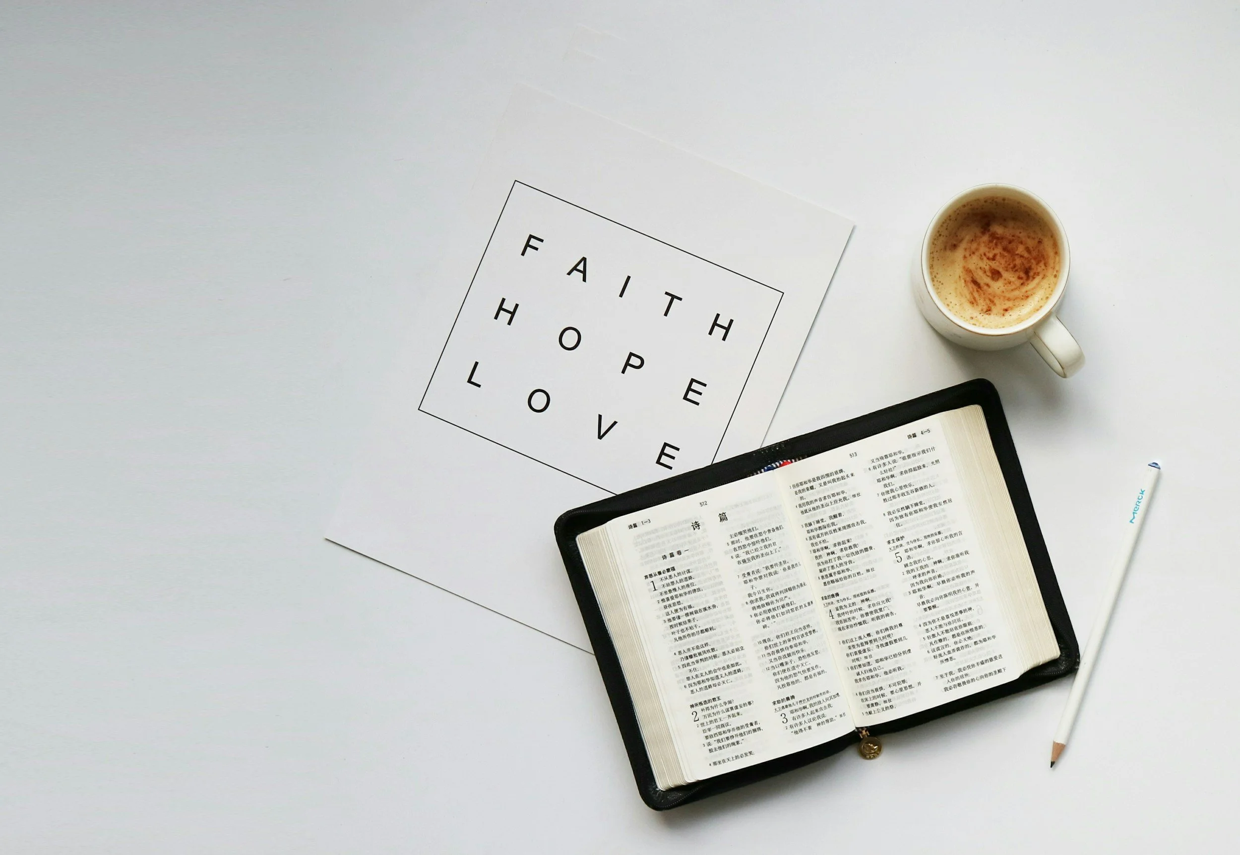 A white desk with an open book displaying text in a foreign language, a tablet, a white mug filled with a frothy beverage, a notepad or letter with the word 'FAITH' arranged vertically in a square grid with the words 'HOPE' and 'LOVE' in the grid, a tube of Mervis product, and a pen.