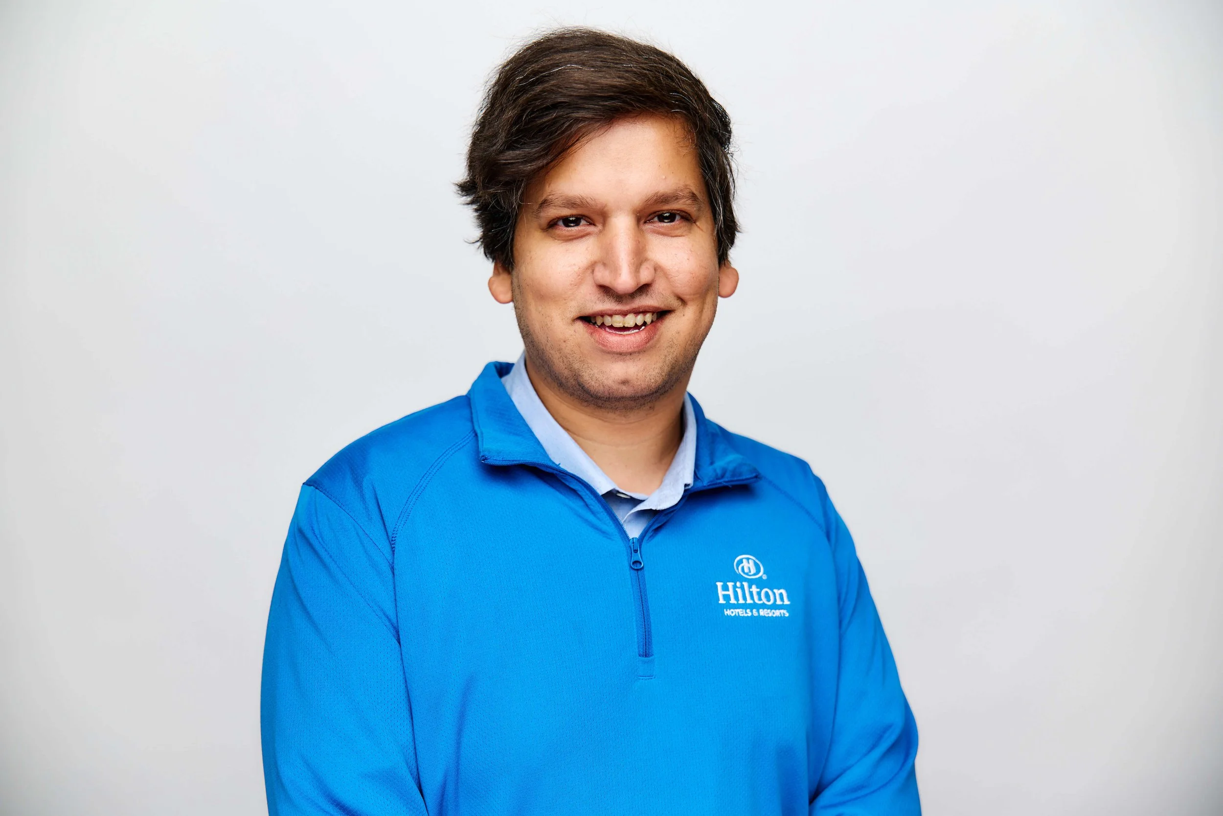 Portrait of a smiling man wearing a blue Hilton Hotels & Resorts jacket against a plain light gray background.