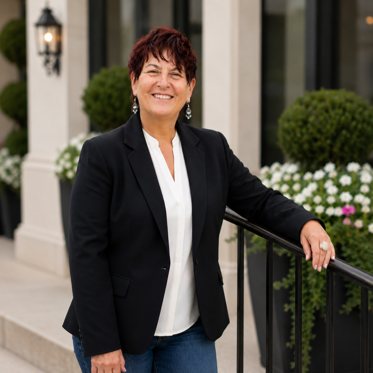 A smiling woman with short, red hair wearing a black blazer and white shirt, standing outdoors with a black railing, green bushes, and white flowers in the background.