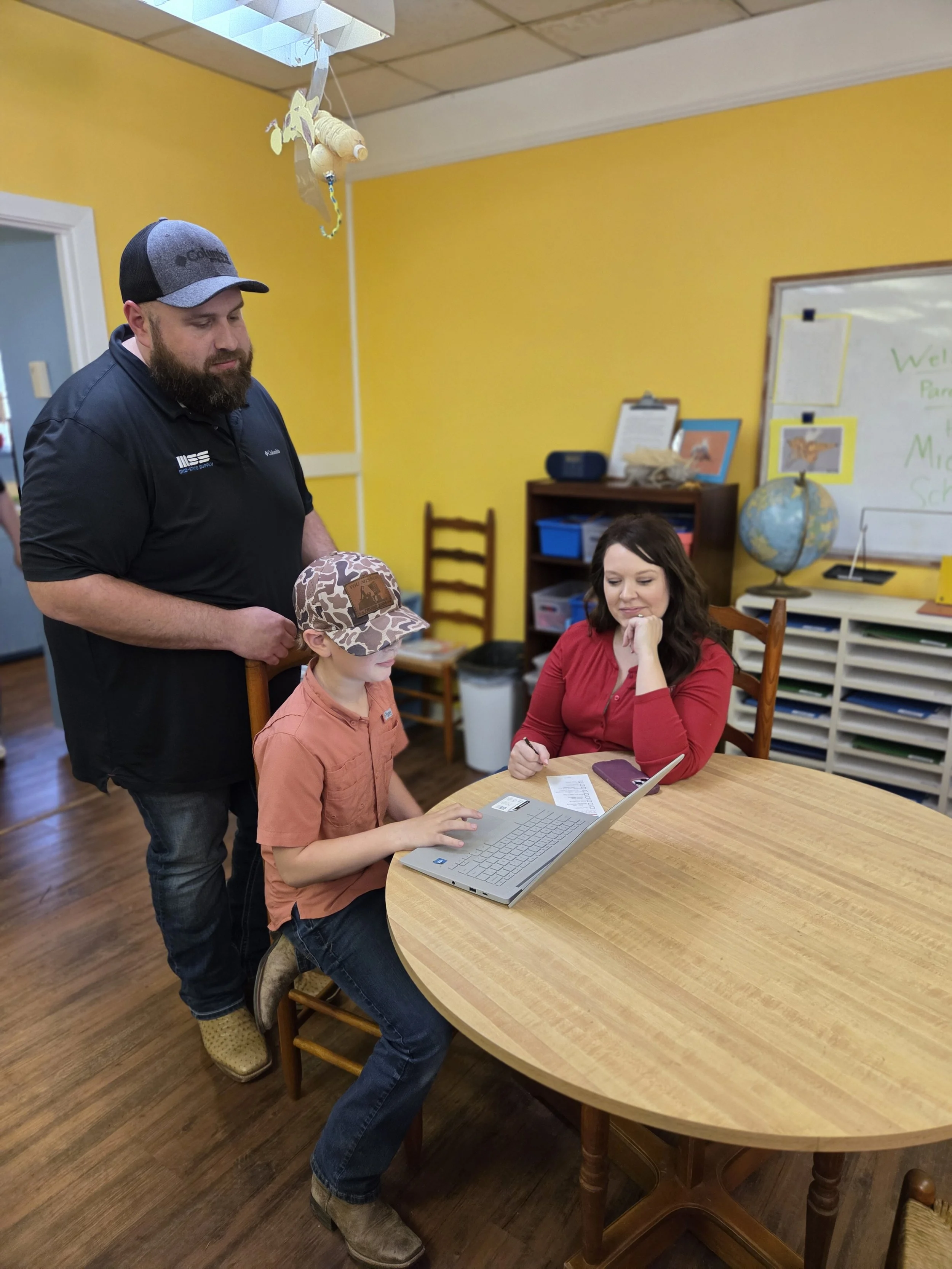 A man, woman, and child are gathered around a wooden table in a classroom. The woman is seated at the table, working on a laptop, while the man and child stand nearby. The woman has dark hair and is wearing a red shirt, the man has a beard and is wearing a dark shirt and a trucker hat, and the child is wearing a tan camouflage hat and an orange shirt. The classroom has yellow walls, a whiteboard with colorful writing, a globe, and a bookshelf in the background.