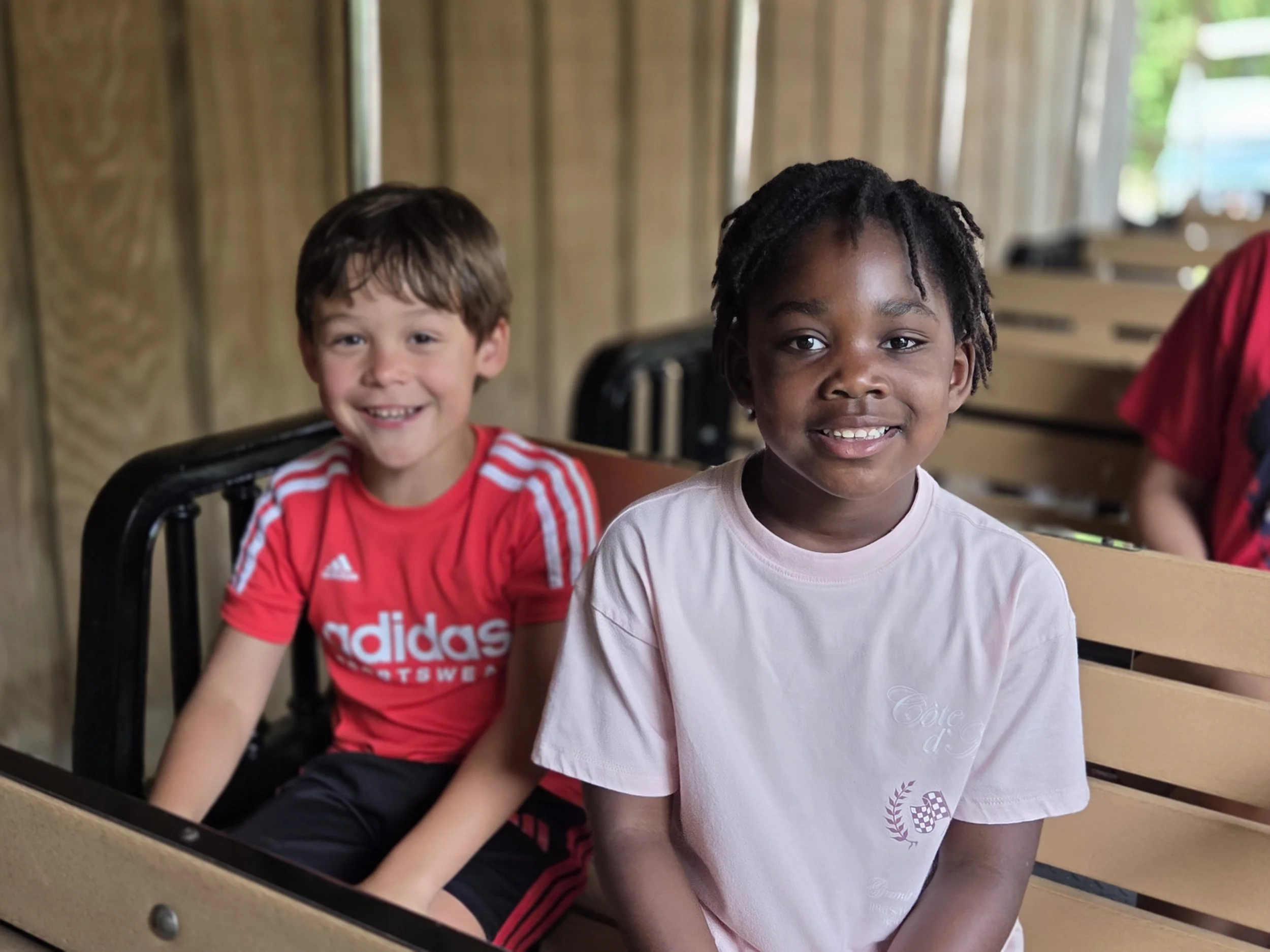 Two boys sitting side by side smiling in a wooden room, one wearing a red Adidas shirt and the other in a light pink shirt with an emblem, with blurred background.