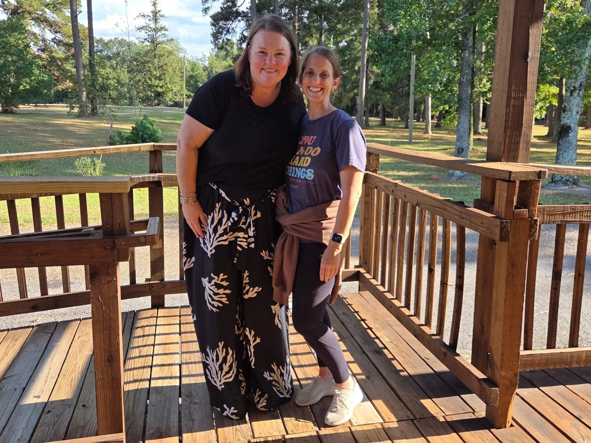 Two women smiling and standing on a wooden deck outdoors in a sunny park-like setting with trees in the background.