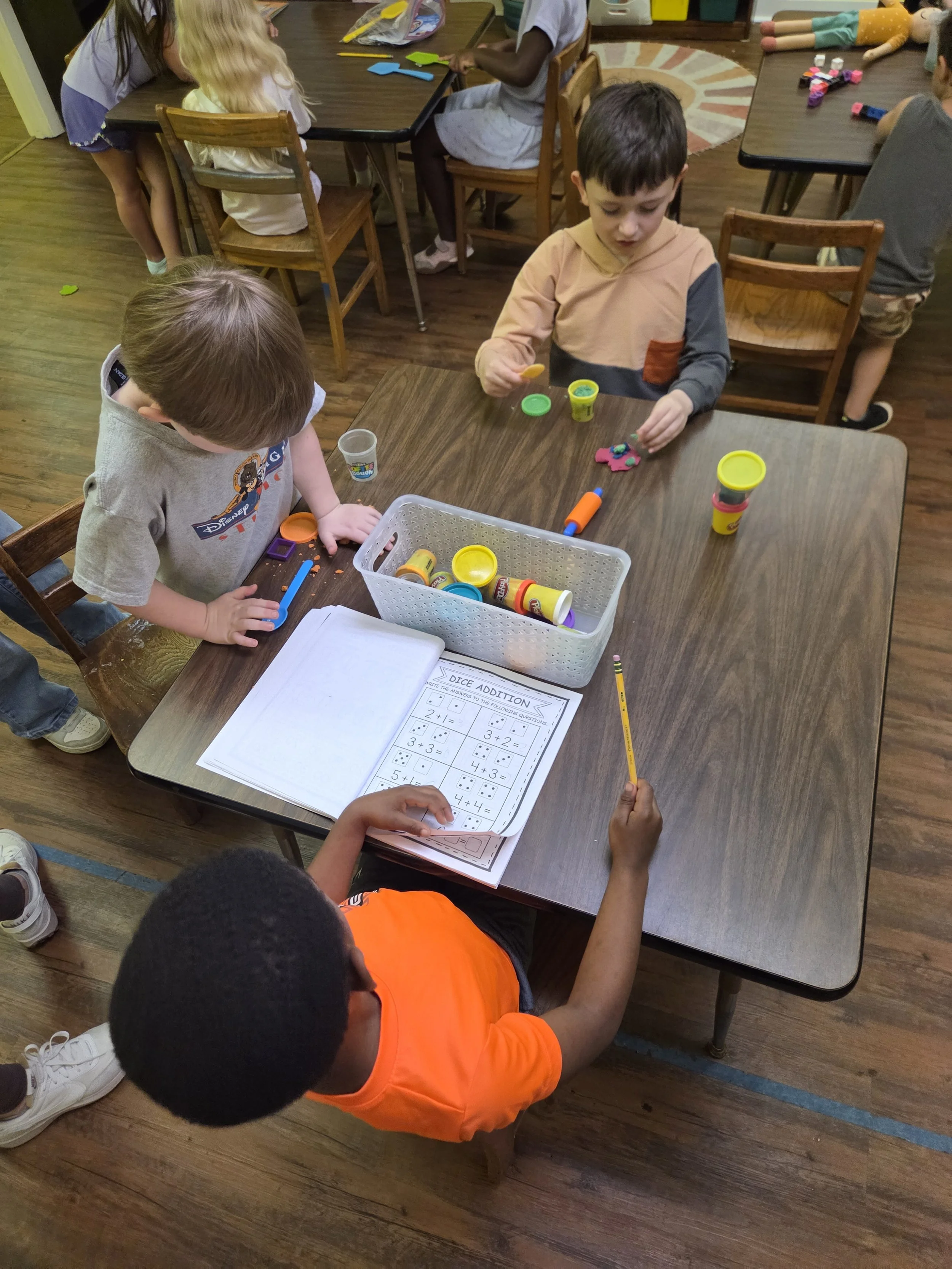 Children doing activities at a classroom table, with Play-Doh, puzzles, and a worksheet on math. Some children are sitting, others are standing or kneeling, engaging with various toys and educational materials.